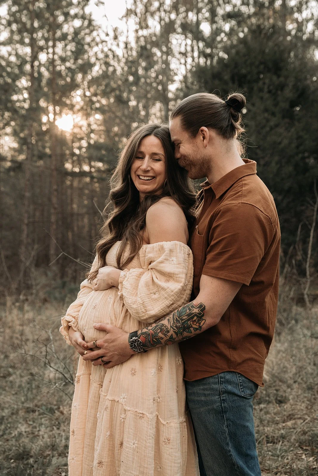 A happy couple, with the woman being pregnant, embracing outdoors during sunset, smiling and touching her belly.