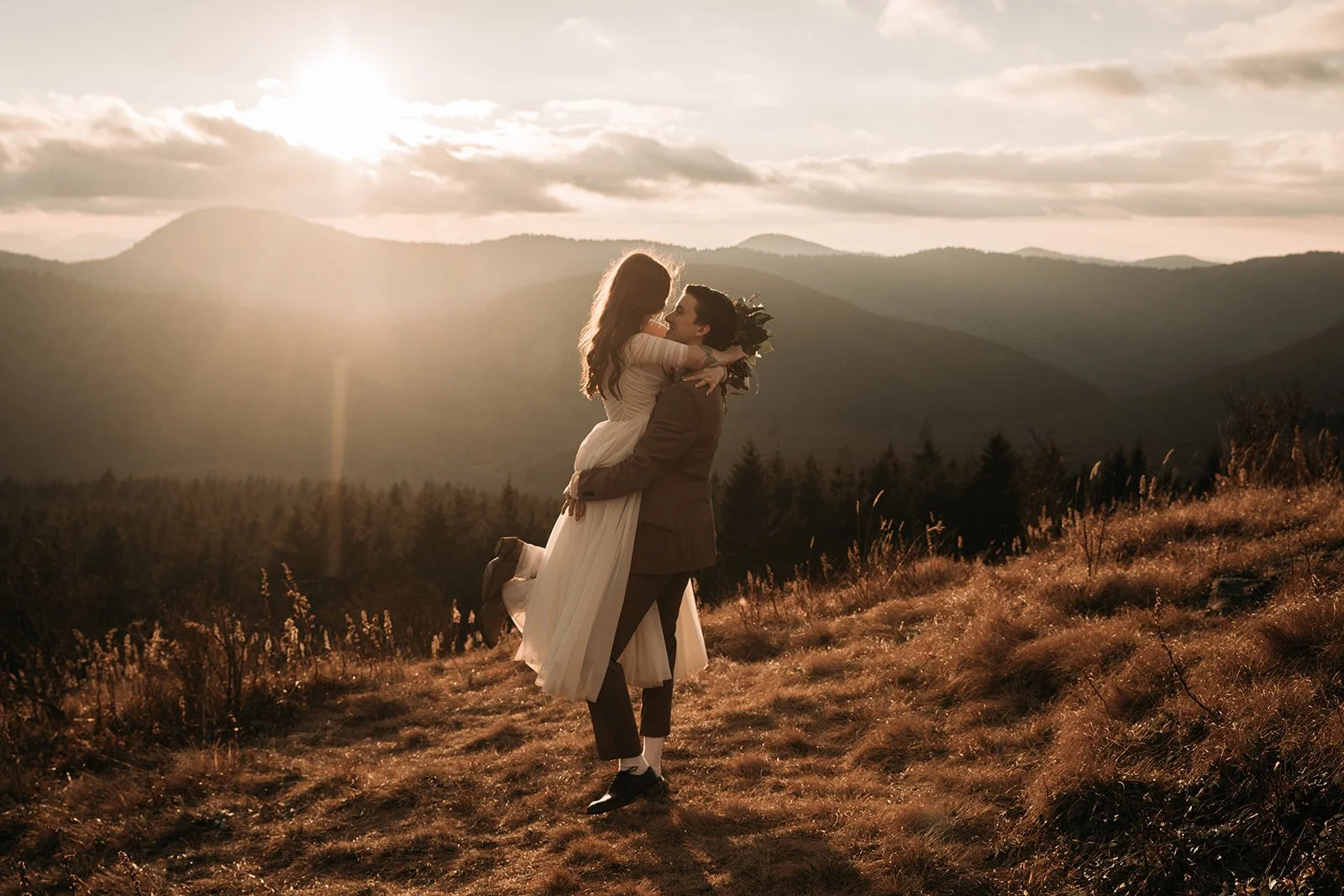 A couple in wedding attire, with the groom holding the bride in his arms, standing outdoors on a grassy hill at sunset with mountains in the background.