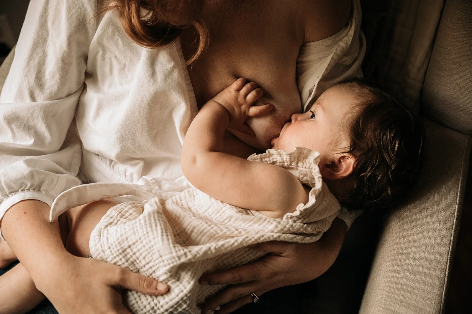 A mother is breastfeeding her young child, who is lying on her chest, on a beige couch. The child is looking at the mother and touching her breast while nursing.