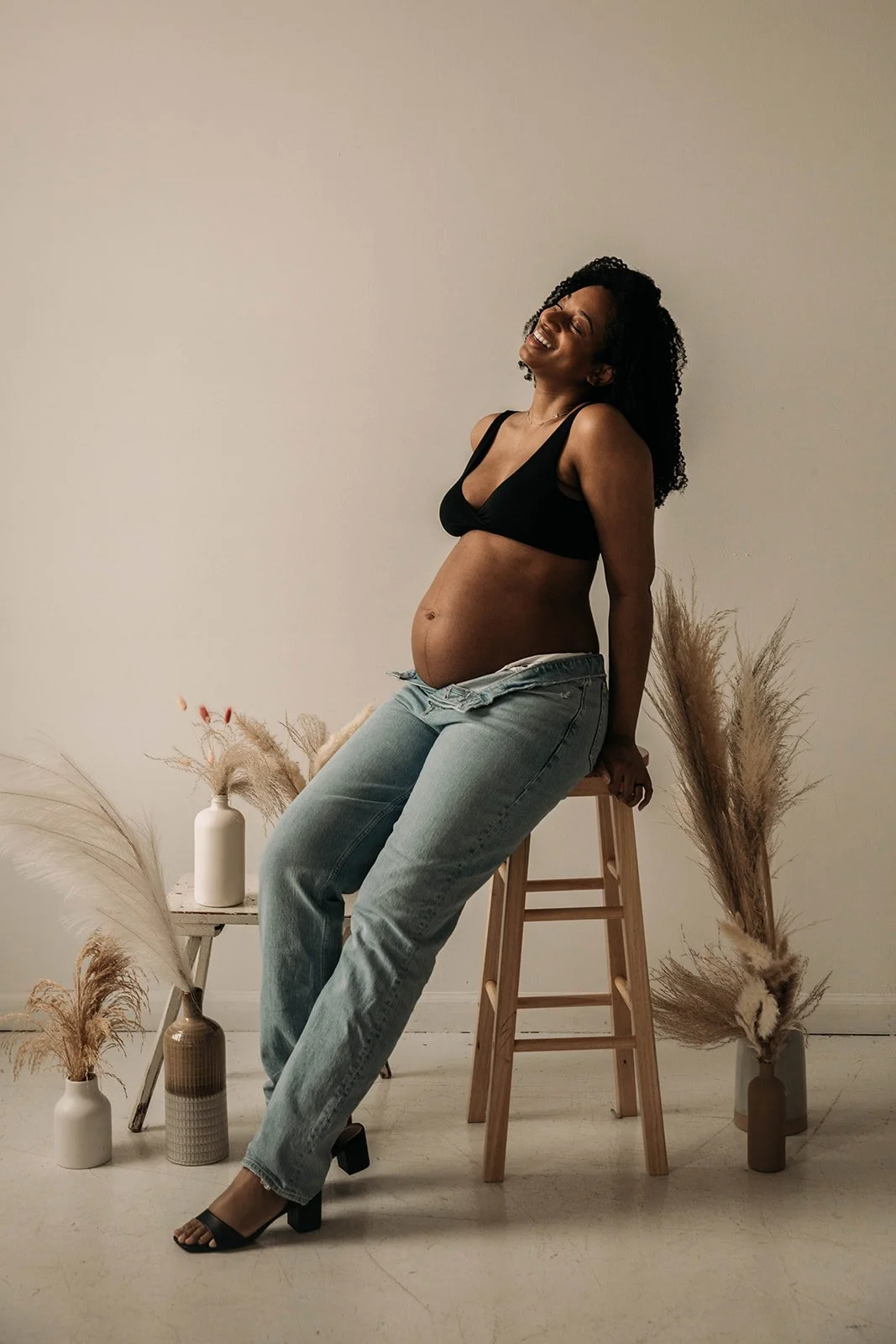 An expecting woman with curly hair, wearing a black crop top, ripped jeans, and black sandals, sitting on a high wooden stool in a minimalist room with beige and white vases filled with pampas grass around her, smiling with eyes closed.