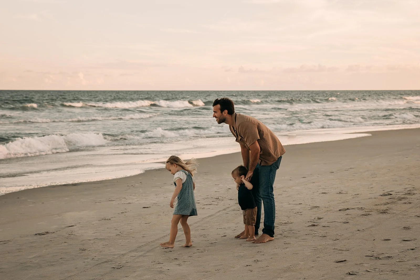 A man and two young children playing on the beach at sunset. The man is standing, holding one child's hand, while the other child walks in front of them near the shoreline.
