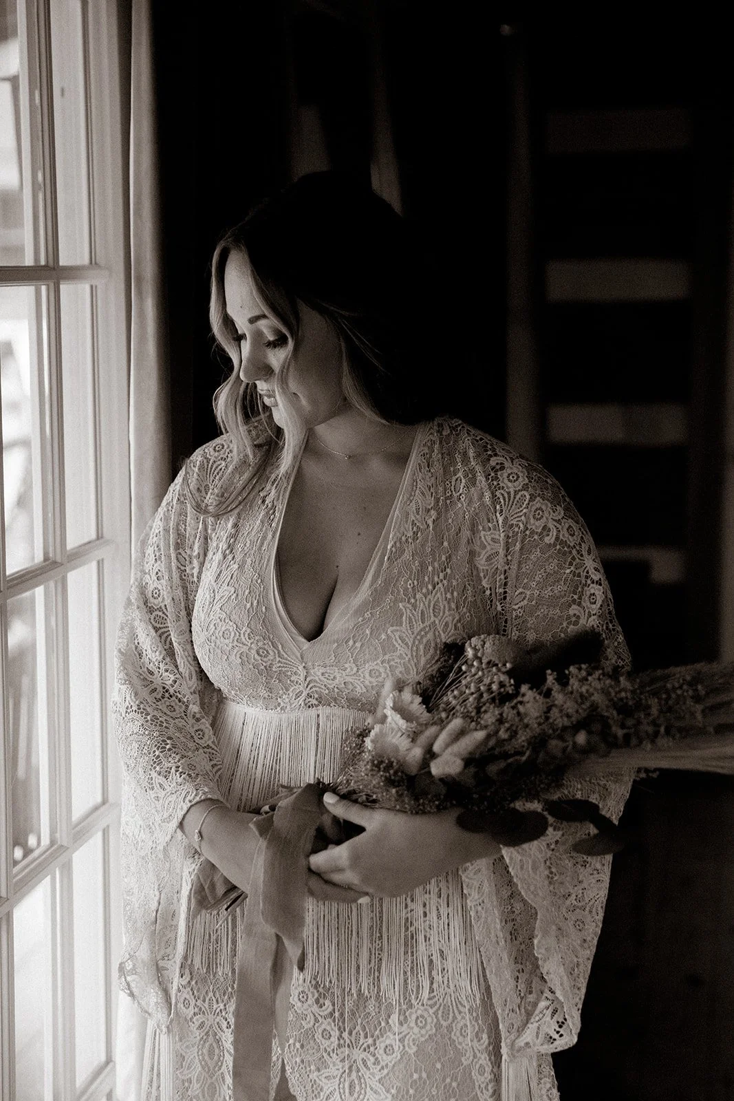 A woman in lace dress holding a bouquet of flowers, standing near a window, softly lit.