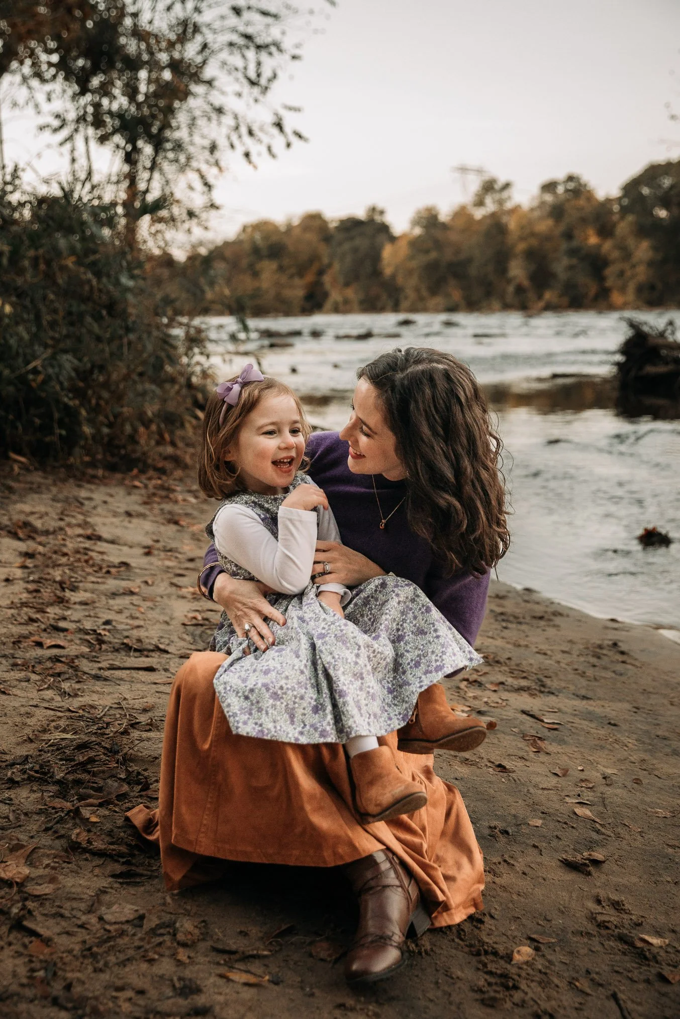 A woman and a young girl sit together on a sandy riverbank surrounded by trees, smiling and enjoying each other's company during a peaceful outdoor moment.