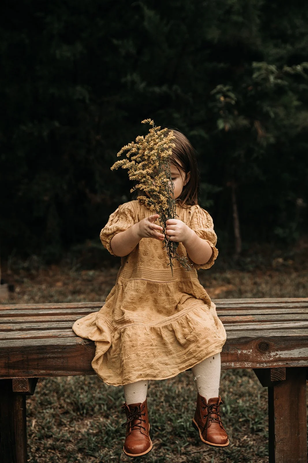 A young girl sitting on a wooden bench outdoors, holding a bunch of dried wildflowers in front of her face, with a dark forest background.