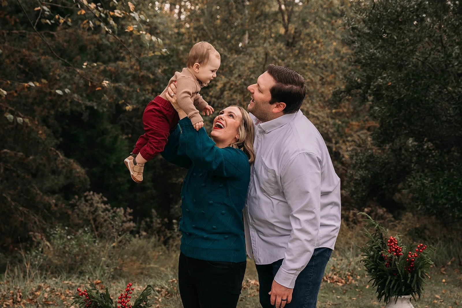 A family of three outdoors, with a woman lifting a young child while a man stands beside them. The background has autumn trees and there is a flower arrangement with red berries on a small table.
