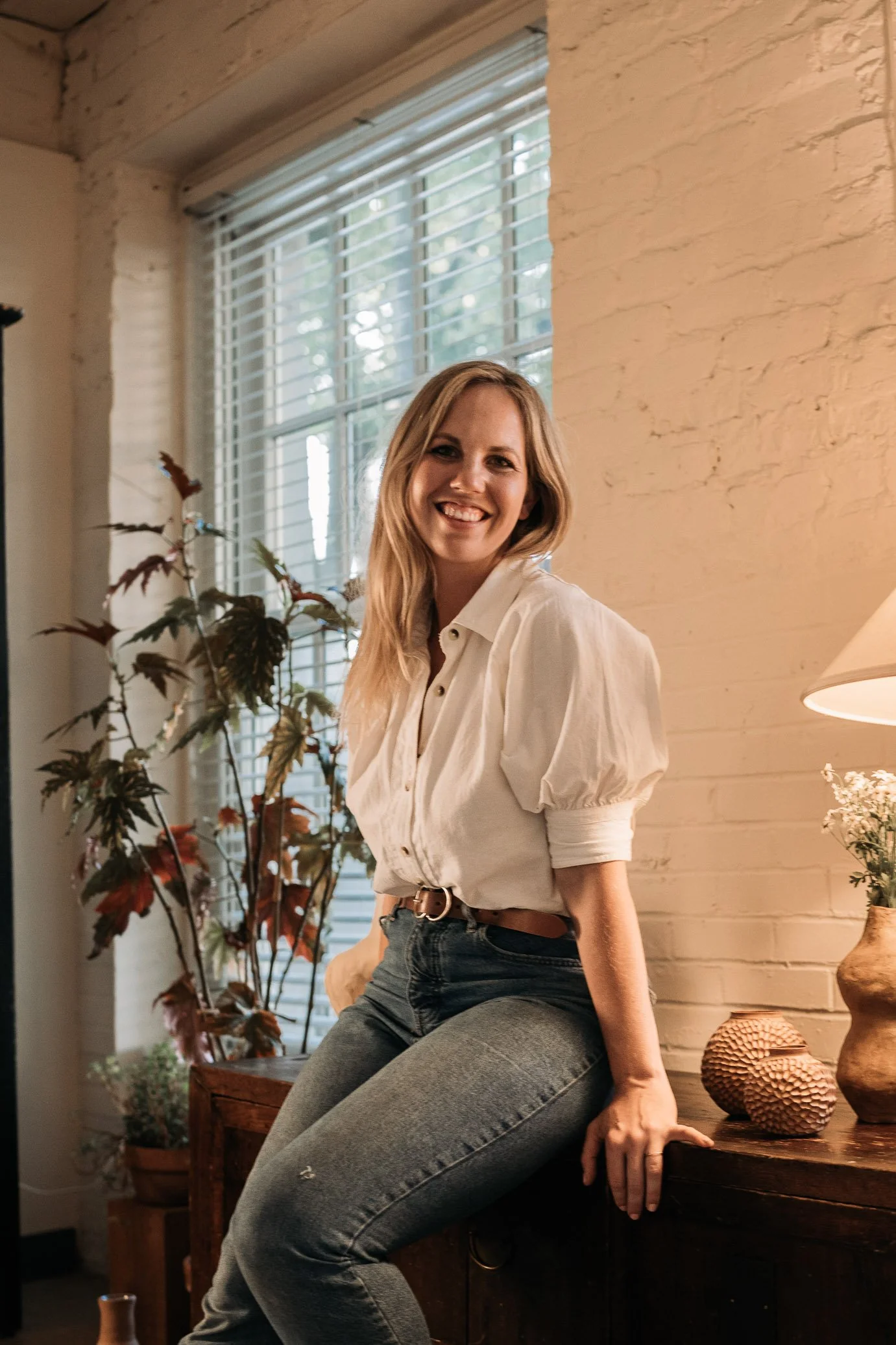 A smiling woman sitting on a wooden cabinet near a window with white blinds, with a plant and decorative vases nearby.