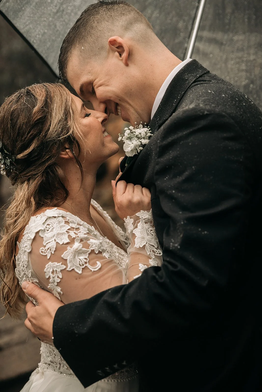 A bride and groom embrace under an umbrella, smiling with their faces close, in a wedding moment.