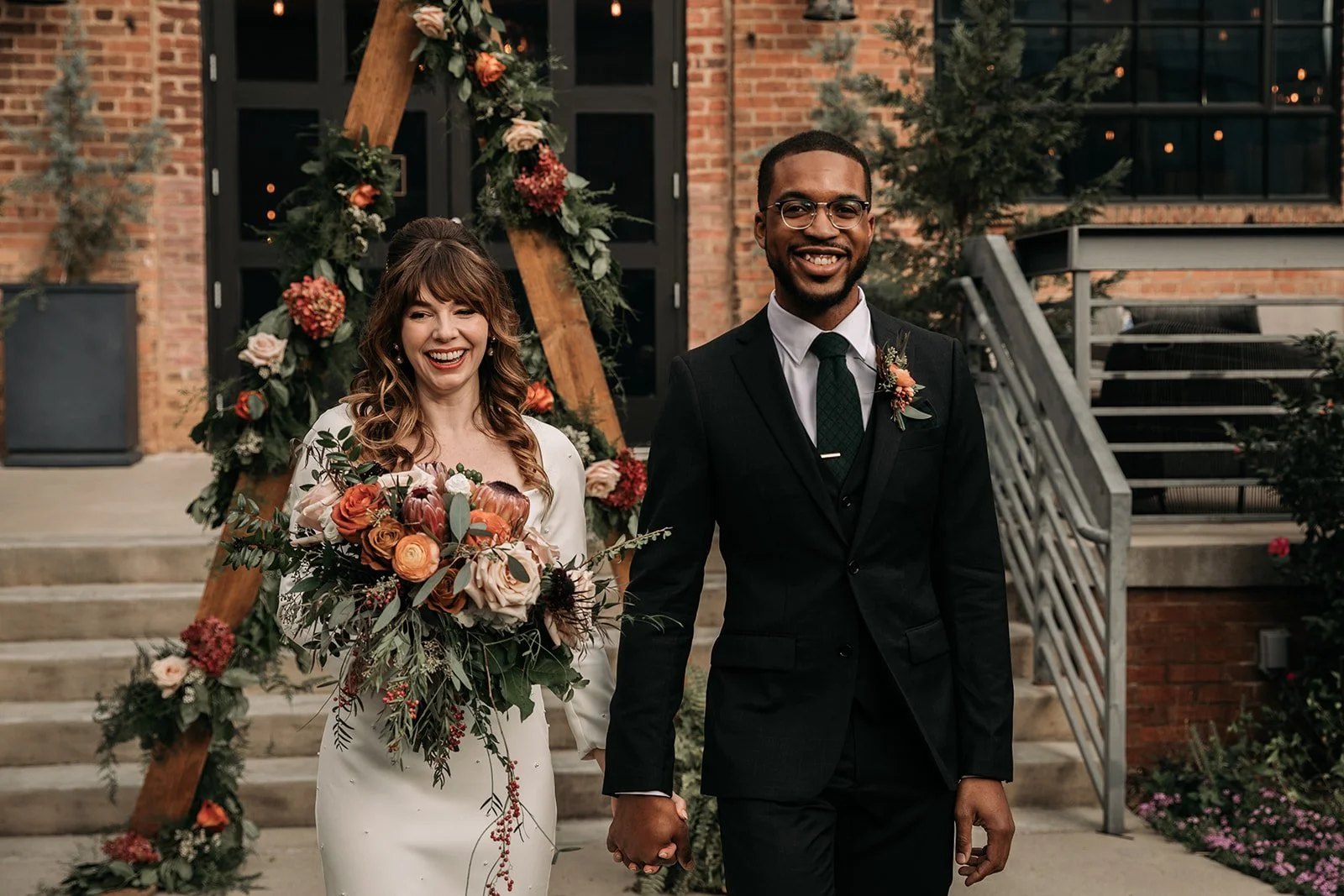 A newlywed couple holding hands and walking out of a wedding venue decorated with flowers and greenery. The bride is wearing a white wedding dress and holding a large bouquet, and the groom is dressed in a black suit with a tie and boutonniere. They are smiling and looking happy.