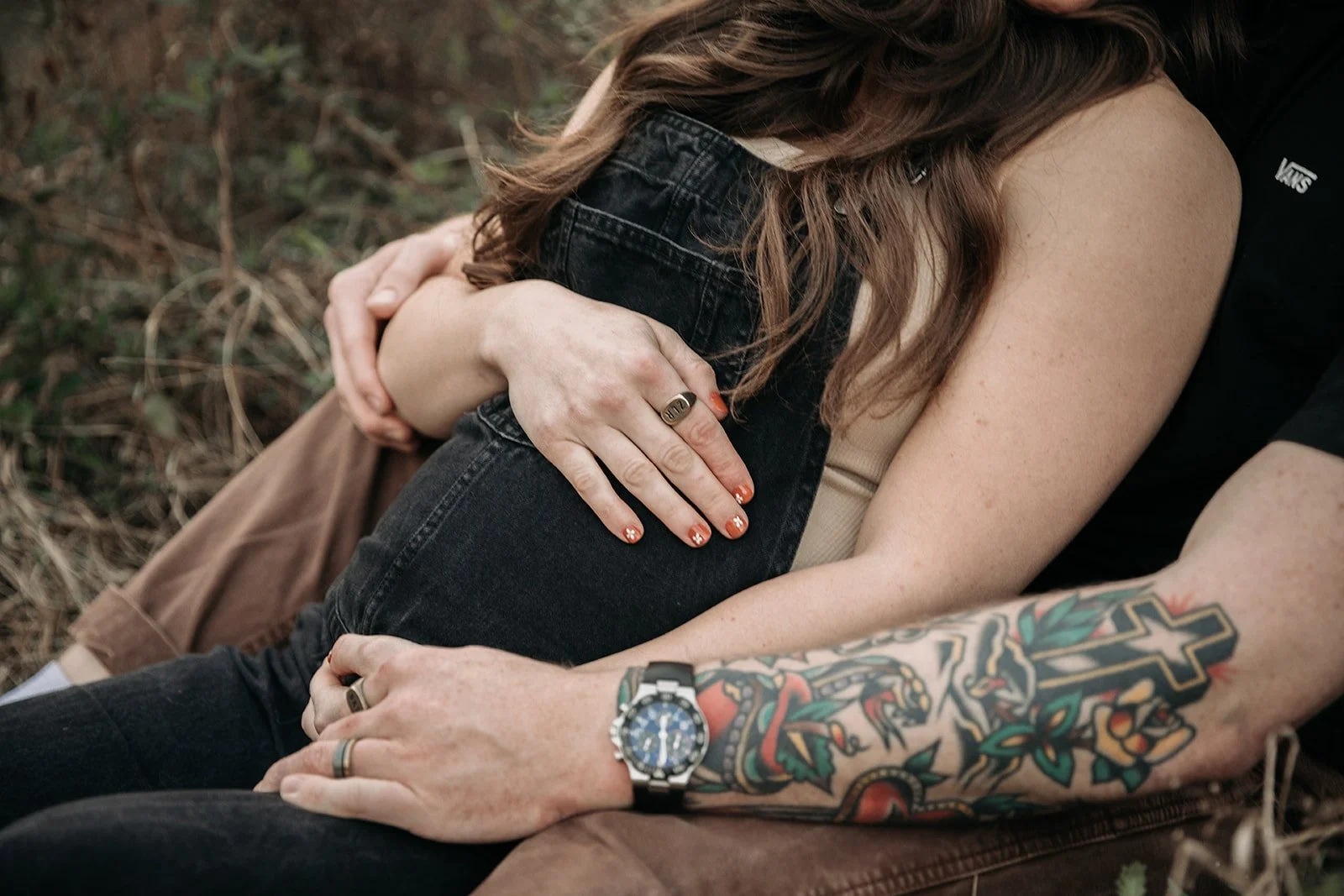 Close-up of a couple sitting on the ground, the woman hugging the man's arm, both with visible tattoos and wearing rings and watch. The woman has long brown hair, and both are dressed casually.
