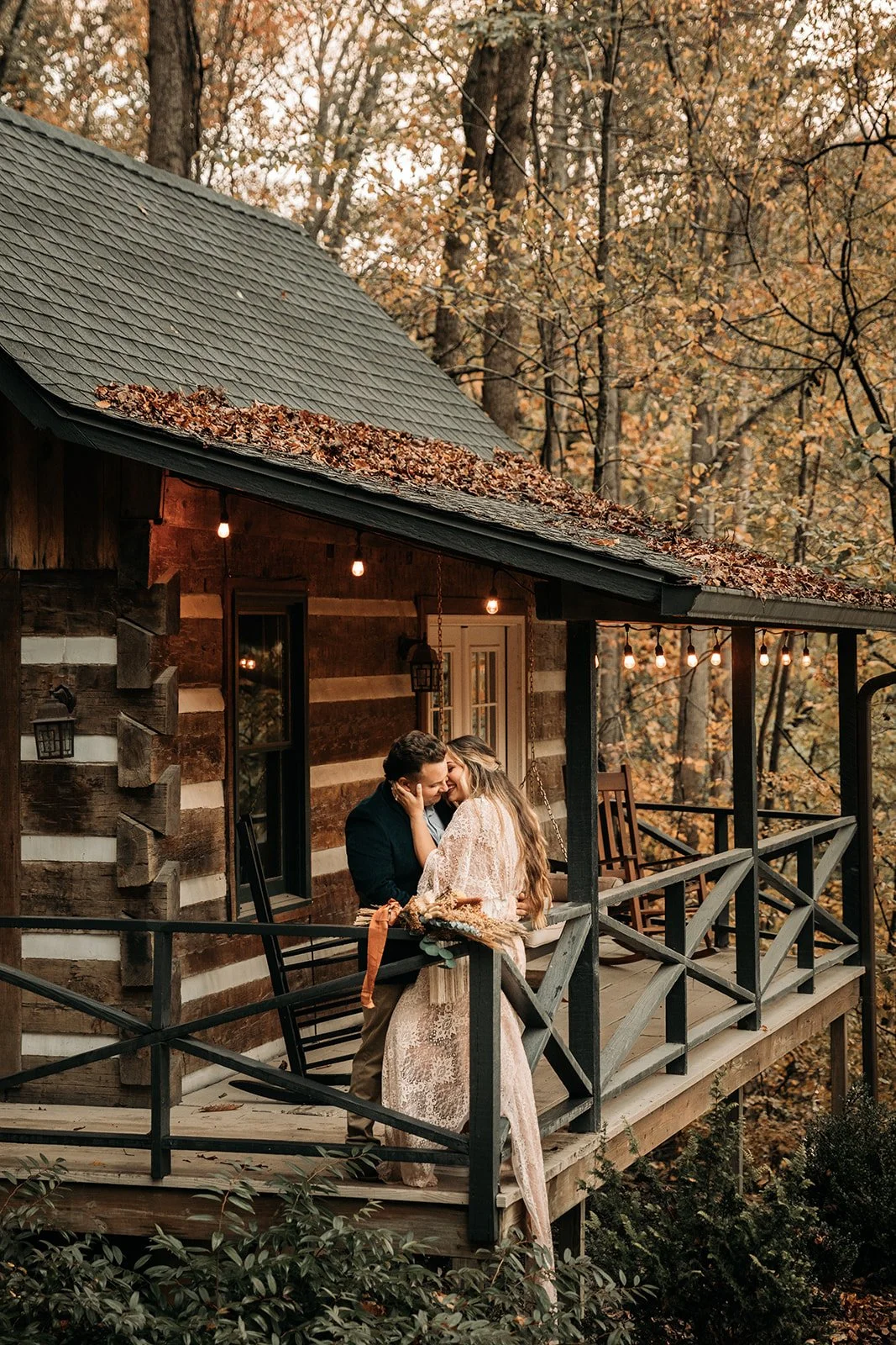 A couple on a wooden porch of a rustic cabin, embracing each other lovingly, surrounded by autumn trees and string lights.