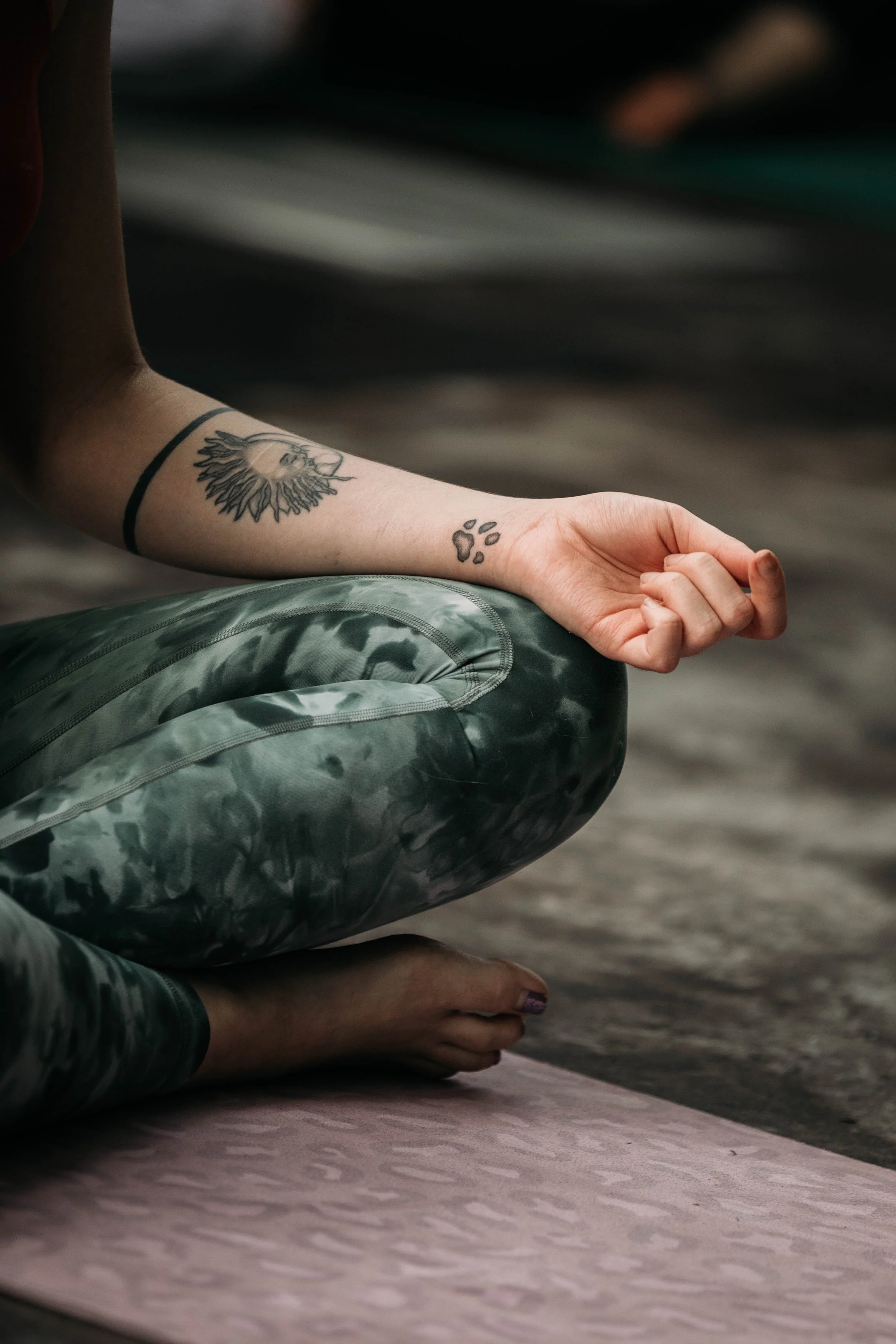 A person sitting cross-legged in a meditation pose on a yoga mat, with one hand resting on the knee and fingers forming a mudra, wearing camouflage pants and displaying tattoos on the arm.