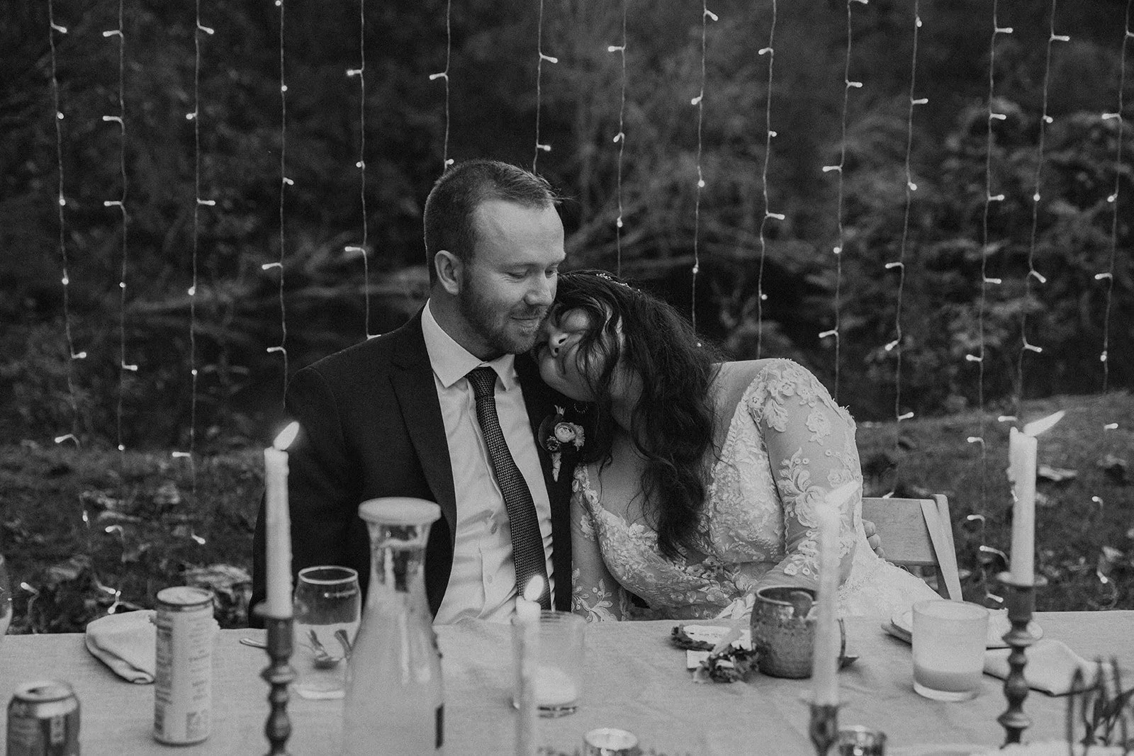 Black and white photo of a couple at a wedding reception, sitting close at a decorated table, with string lights in the background. The woman is resting her head on the man's shoulder, both smiling.