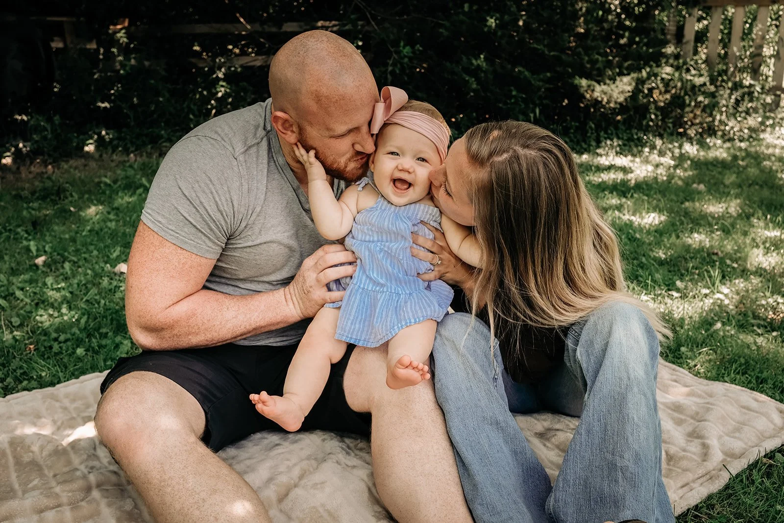 A family of three (father, mother, and baby girl) sitting on a blanket outdoors, sharing kisses and smiling at each other.