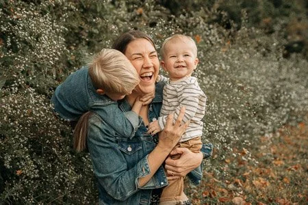 A woman and two children smiling outdoors among bushes and flowers.