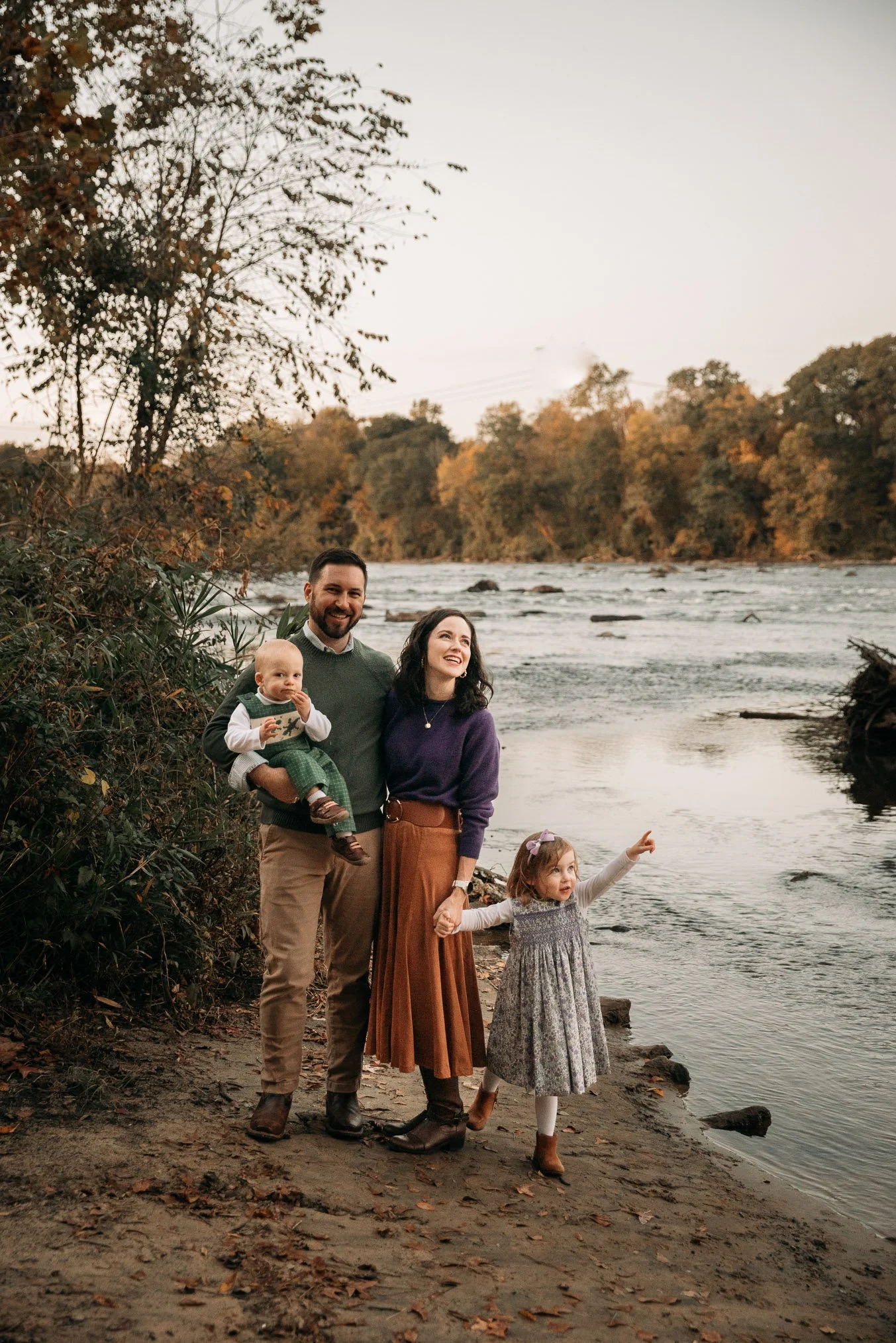 A family of four standing on a riverbank with autumn trees in the background. The man is holding a baby, and a woman and a young girl are holding hands, with the girl pointing towards the river.