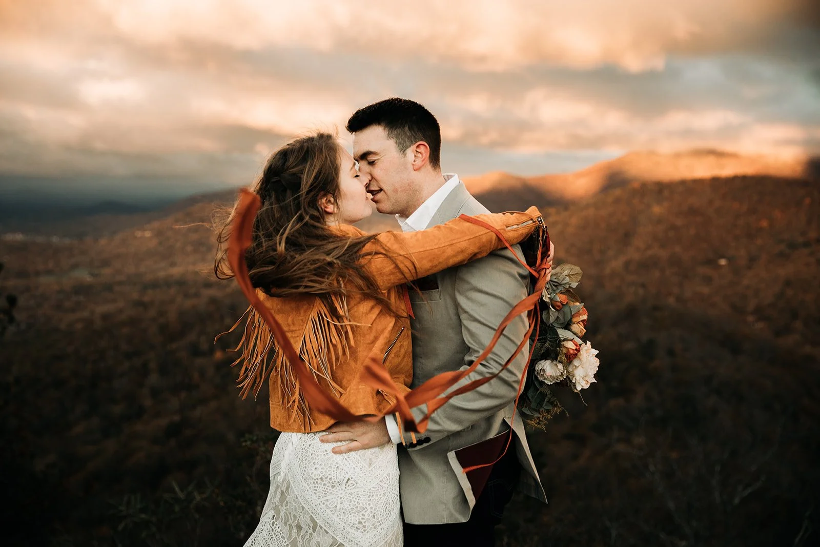 A couple embracing on a mountain during sunset, with the woman in a white lace dress and brown leather jacket, and the man in a gray suit, holding a bouquet of flowers.