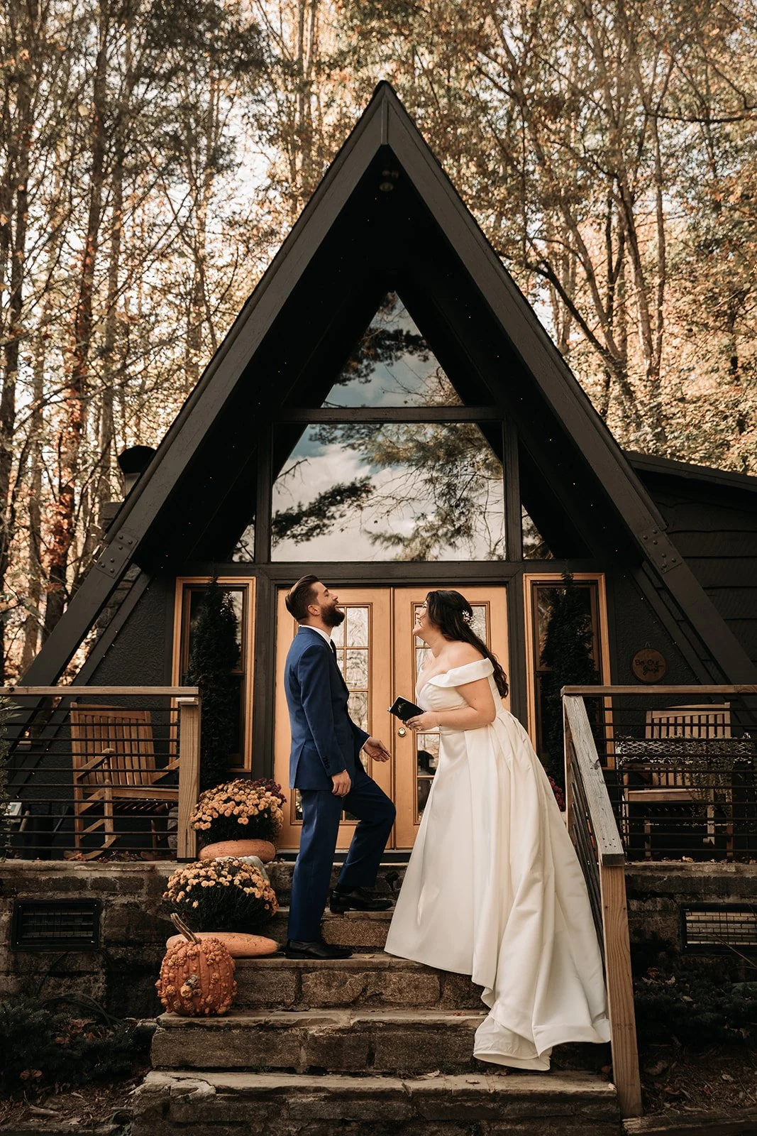 A bride and groom standing on steps outside an A-frame house, facing each other and smiling, during sunset or late afternoon surrounded by trees.