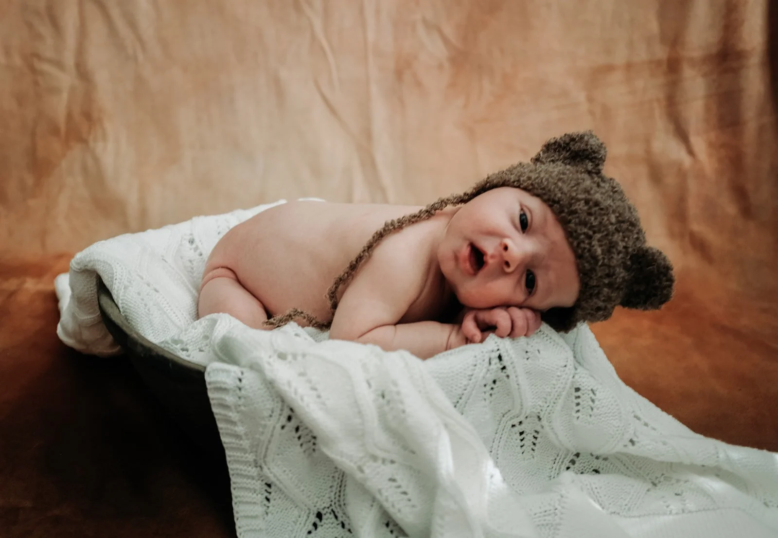 A baby lying on a white knitted blanket, wearing a brown bear hat with ears, on a wooden surface.