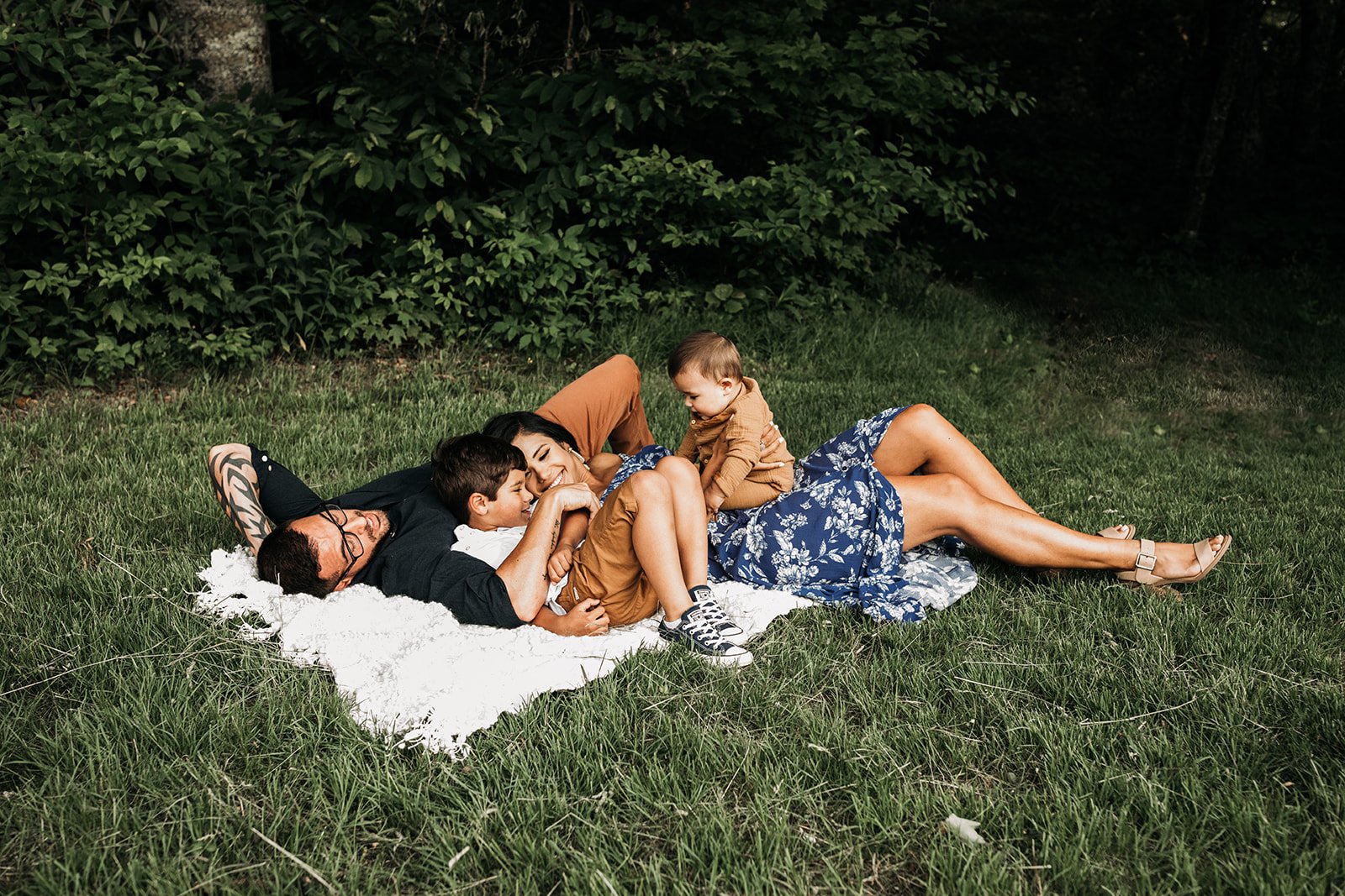 Family of four lying on a white blanket on the grass near bushes, smiling and playing together outdoors.