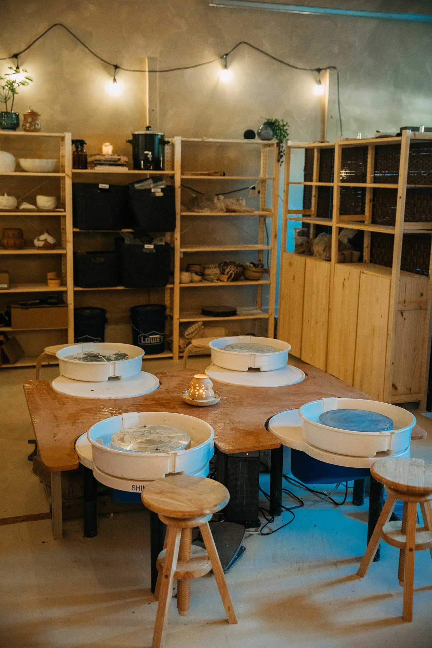 A pottery studio with a large wooden table holding four pottery wheels, surrounded by three wooden stools, with shelves filled with pottery and supplies, string lights overhead, and a candle in the center of the table.