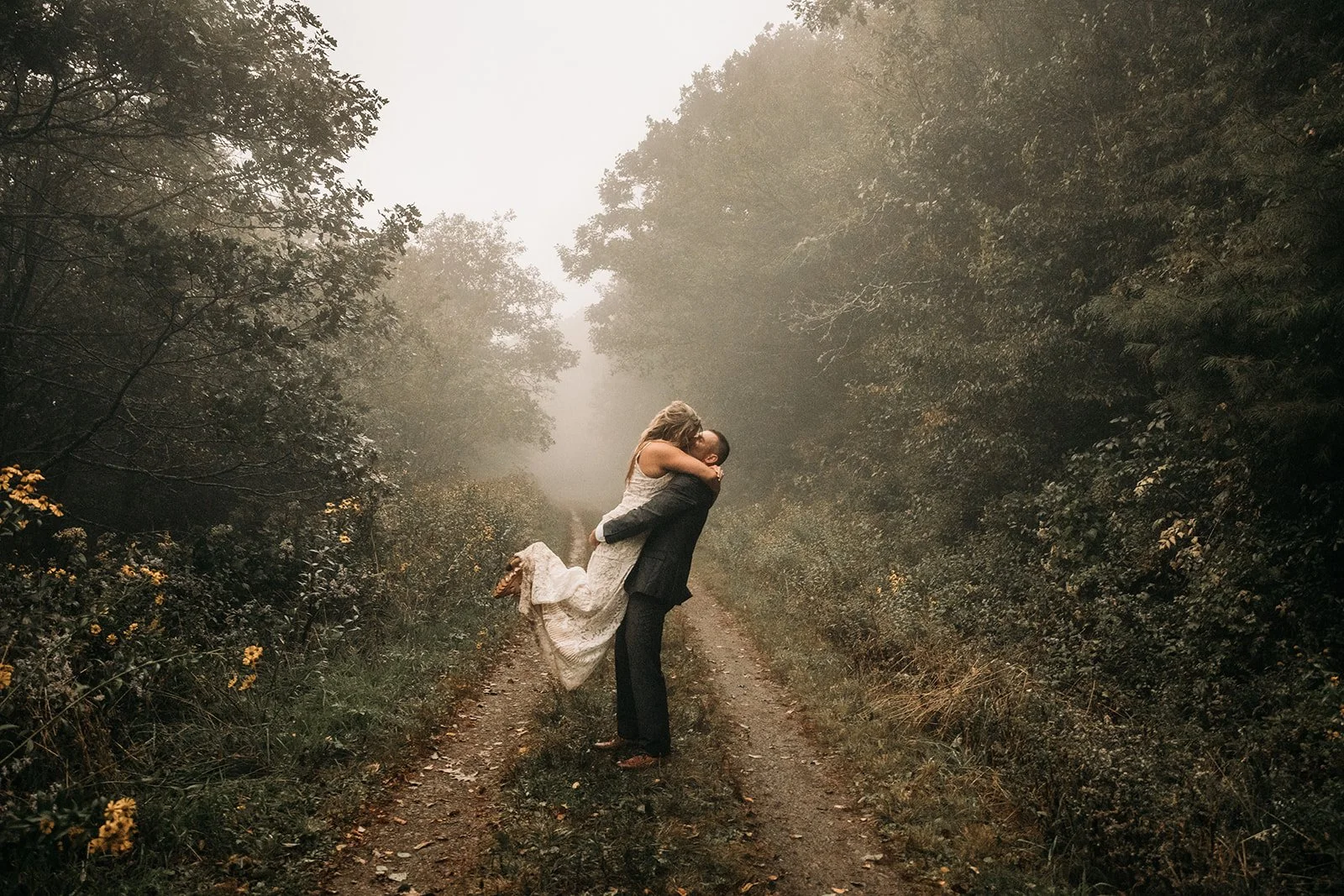 A man in a suit lifting a woman in a white dress in a forested dirt path, misty atmosphere.