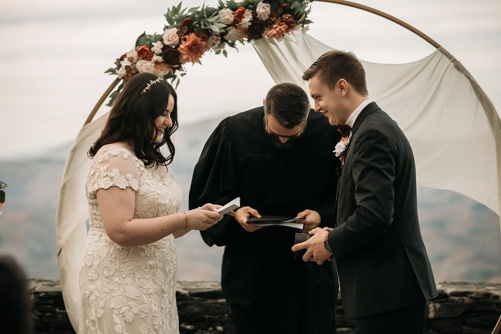 A couple gets married outdoors during the daytime, standing under a floral arch with greenery and pink and white flowers, with a scenic view in the background, as the officiant conducts the ceremony.
