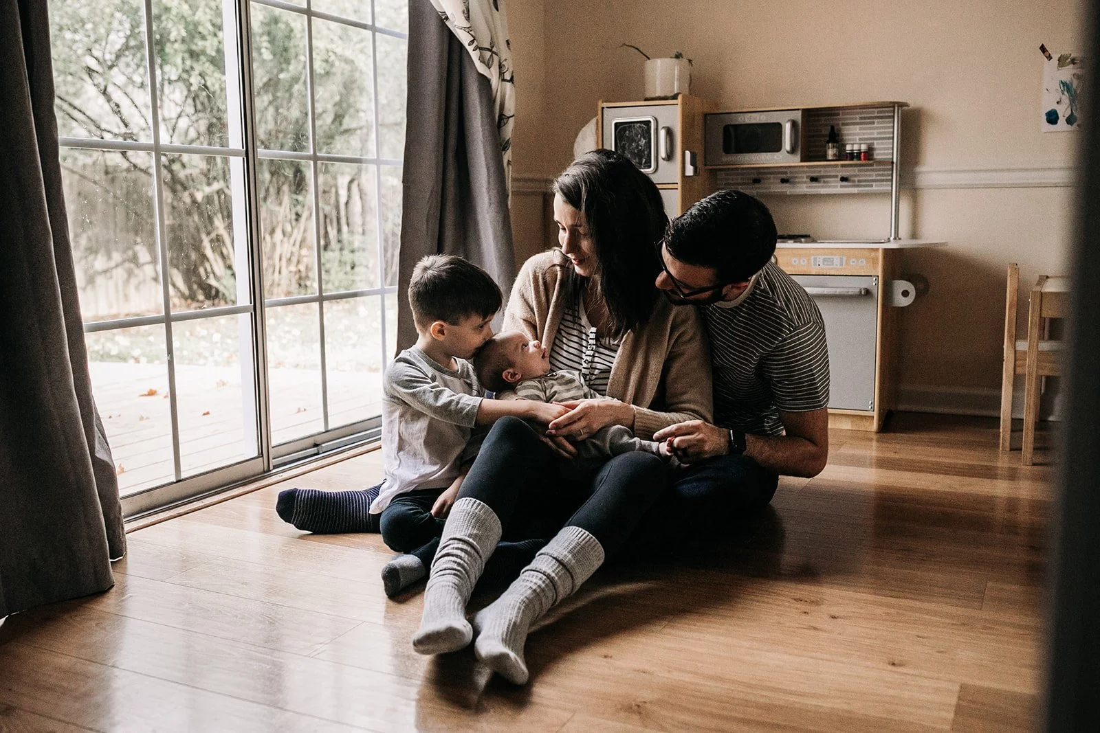 Family of four sitting on the wooden floor inside a living room near sliding glass doors, with trees and a patio outside. The woman is holding a newborn baby, a man and an older boy are leaning close to her, engaging with the baby.