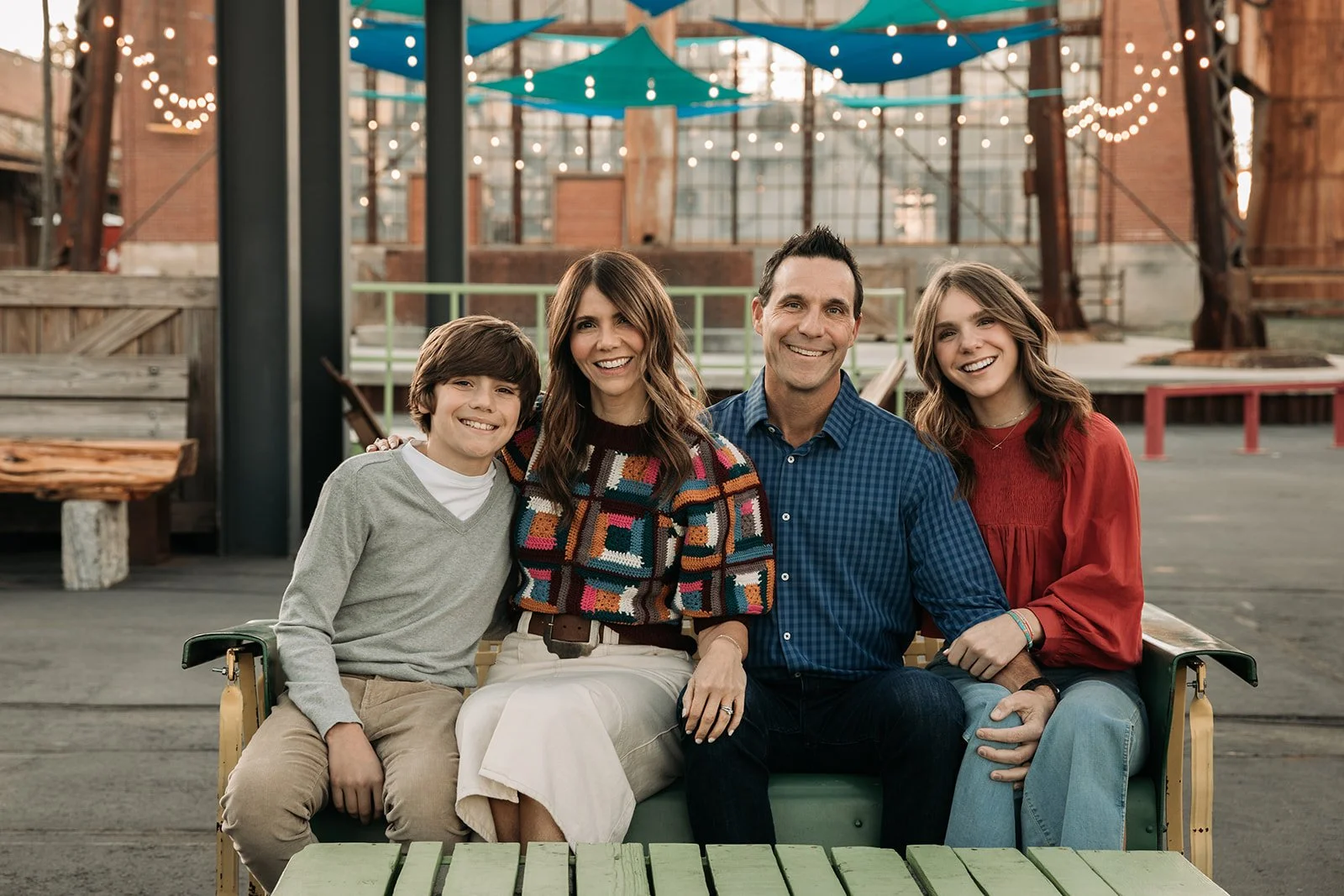 A family of four sitting on a green bench inside a rustic indoor space with string lights and blue ceiling decorations, smiling at the camera.