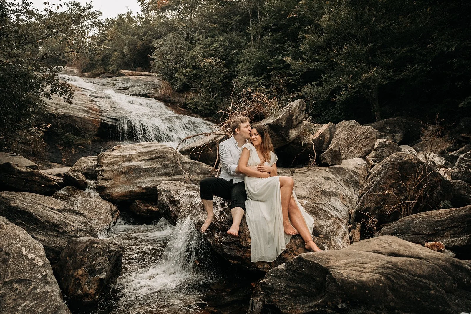 A couple sitting on rocks in a stream with a small waterfall in the background, surrounded by trees.
