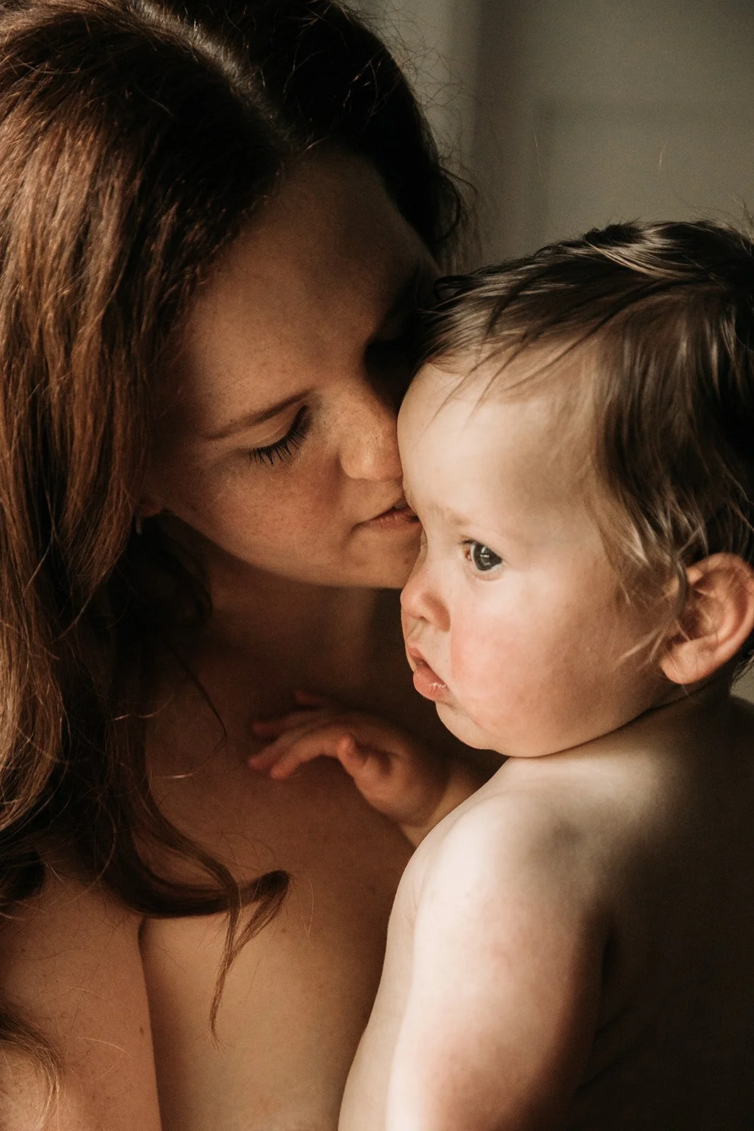 A woman with long brown hair gently kisses a young child with wet hair on the forehead; they are close-up with a neutral background.