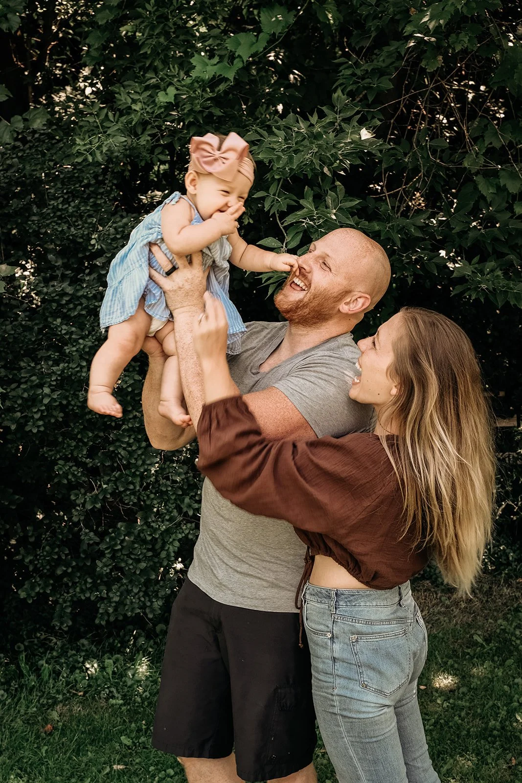 A family of three, parents and their toddler girl, playing outdoors in front of green shrubbery. The father is lifting the girl, who is laughing and touching his nose, while the mother looks on smiling.