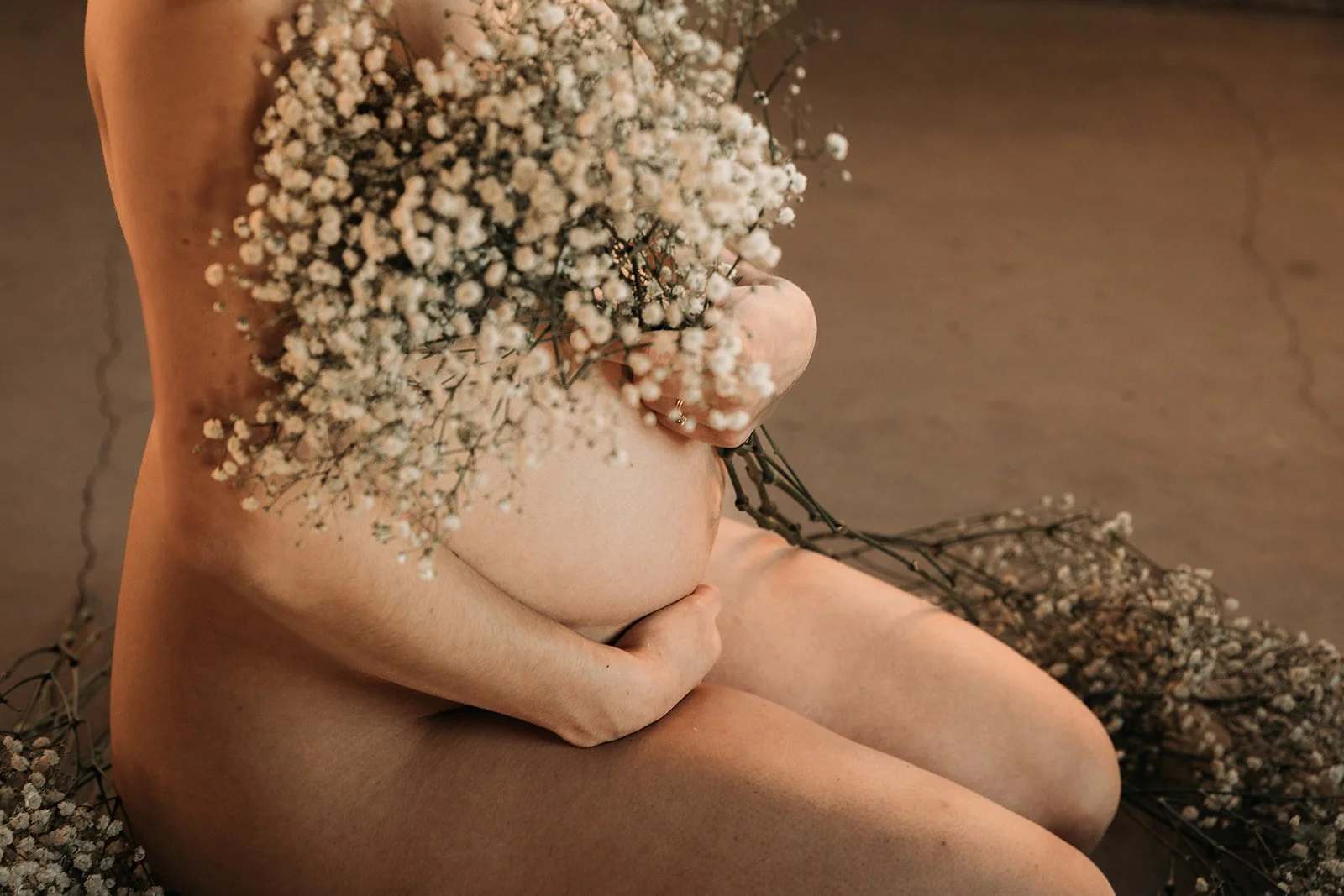 A nude person sitting on a surface, holding a bouquet of small white flowers, with more flowers around them.