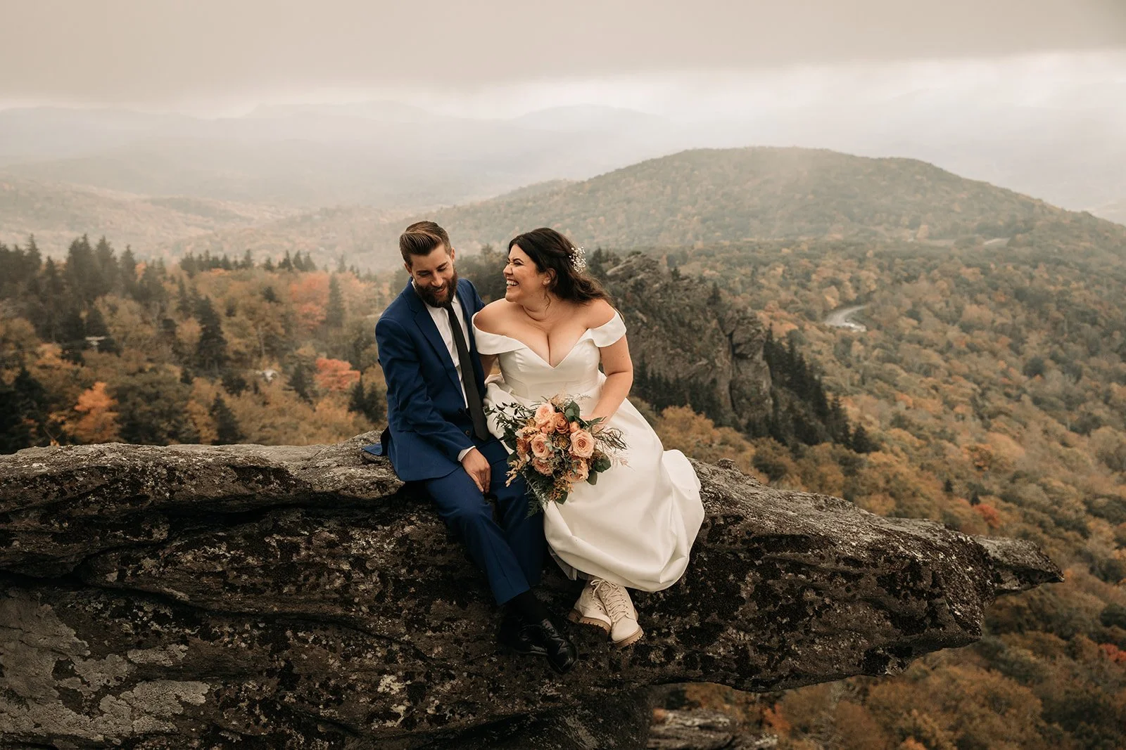 A joyful bride and groom sit on a rocky ledge overlooking a forested mountain landscape, with the bride holding a bouquet of flowers and both dressed in wedding attire.