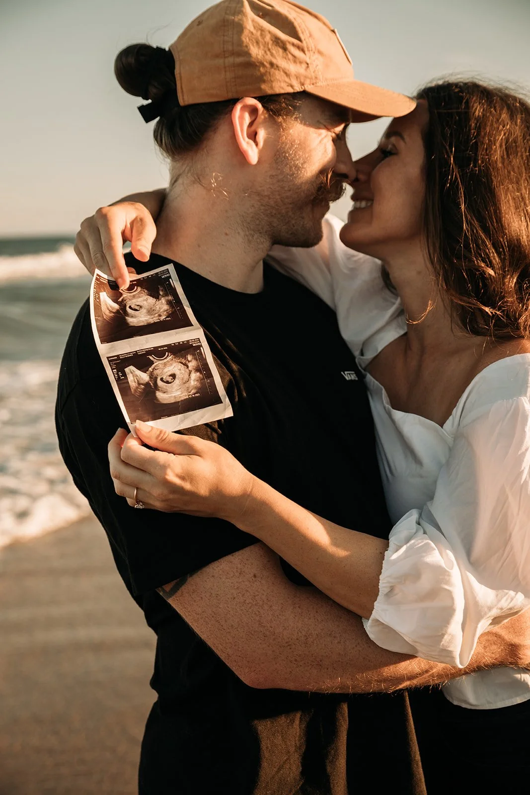 A couple at the beach, holding ultrasound photos and about to kiss with their noses touching, celebrating a pregnancy.
