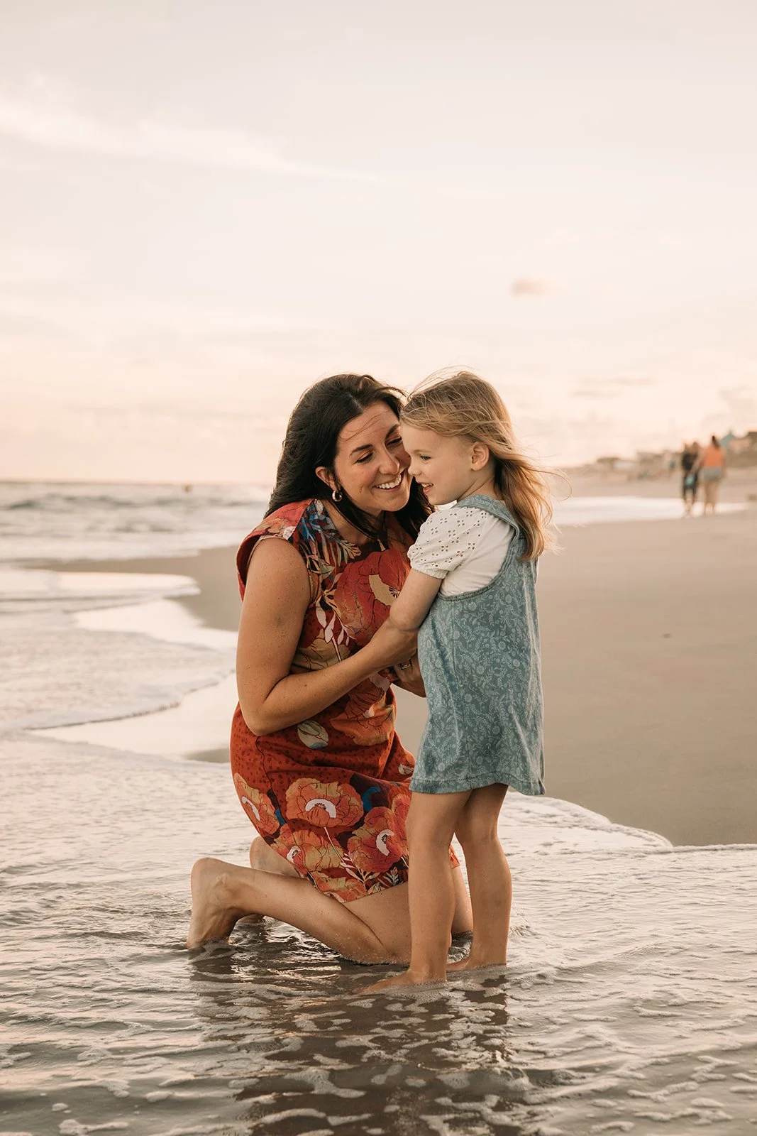 A woman and a young girl are smiling and cuddling on the beach at sunset, with the ocean waves washing around them.
