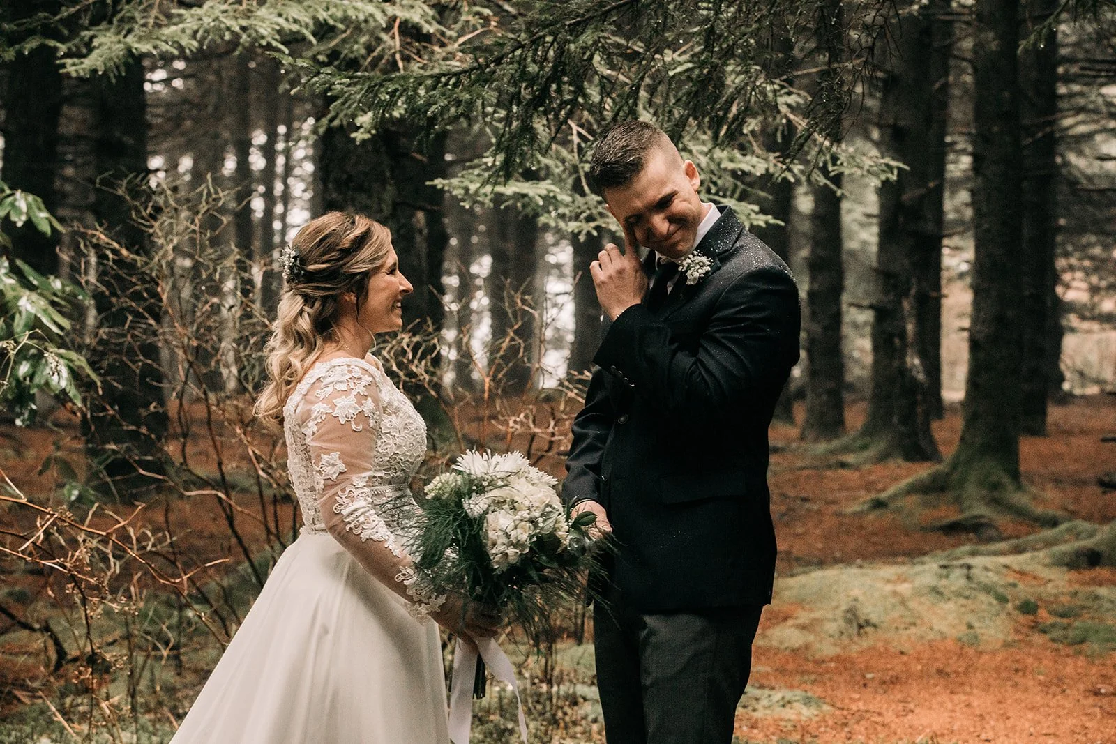 A bride and groom sharing an emotional moment in a forest, with the bride holding a bouquet and the groom smiling as he brushes his hair.