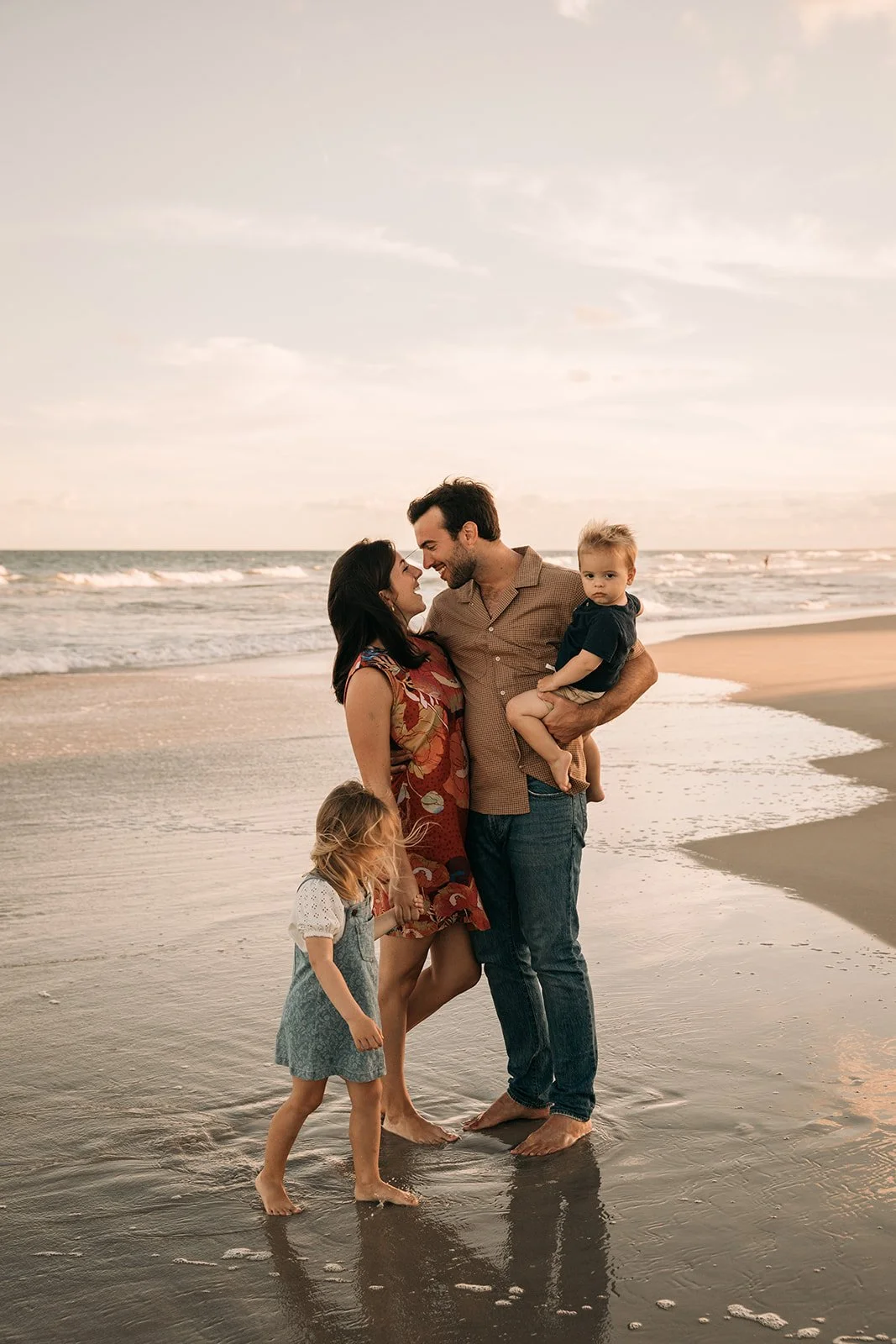 A family of four stands on the beach near the water at sunset, with the parents smiling at each other, the father holding a young boy, and a young girl holding the mother's hand.