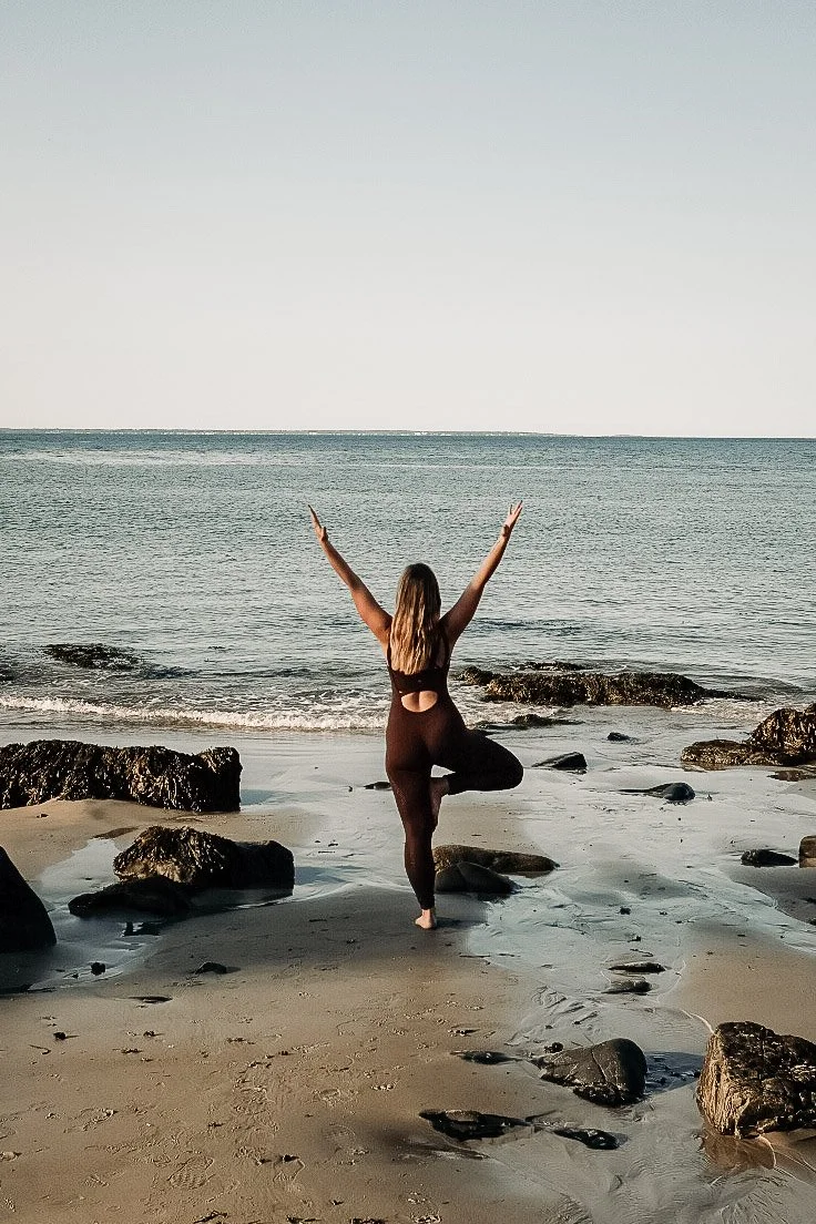 A woman practicing yoga on a beach, standing on one leg with her arms raised, facing the ocean.