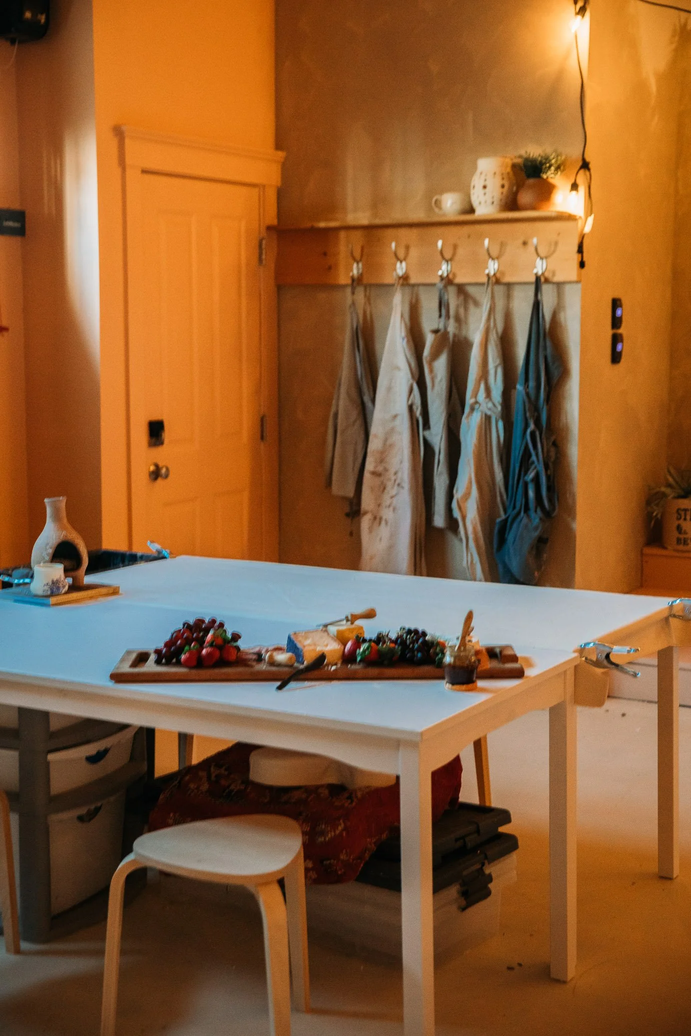 Kitchen with a white table holding a cheese platter, grapes, strawberries, and jam. Coats and aprons hang on hooks on a wooden wall. Warm lighting with a hanging bulb and potted plants on a shelf.