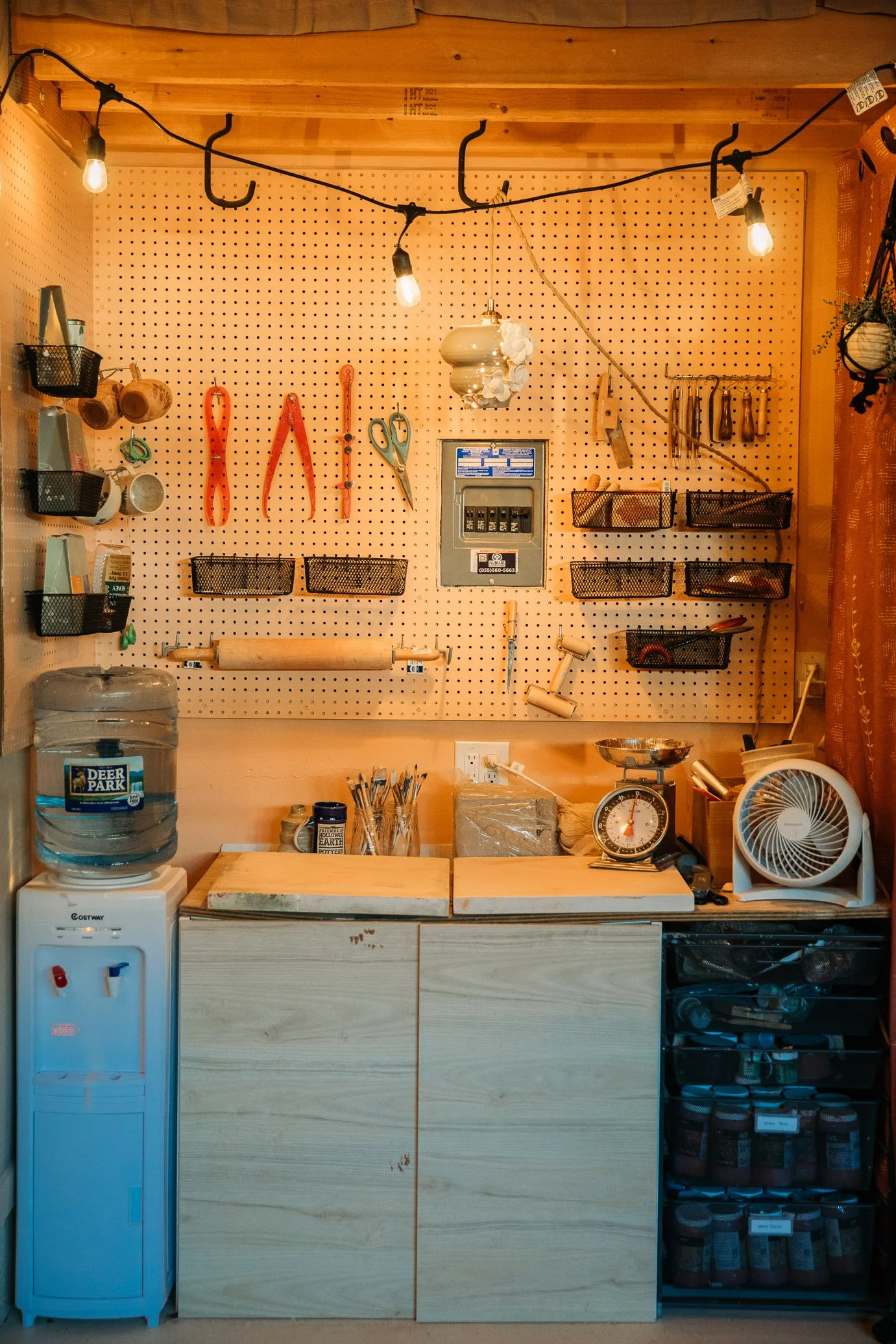 A cozy workshop with pegboard wall holding tools such as scissors, pliers, and knives, alongside baskets, a scale, and kitchenware. A water dispenser, fan, and work surfaces are visible below.