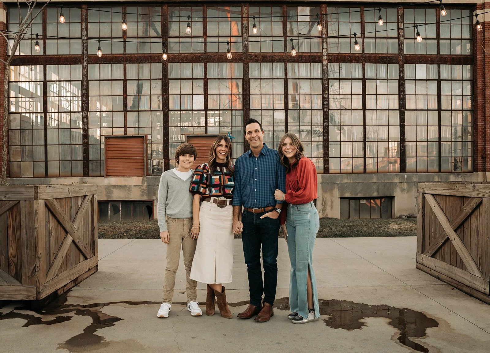 A family of four standing outdoors in front of a large industrial-style glass building with string lights. The family members are smiling and holding hands, including a young boy, two women, and a man.