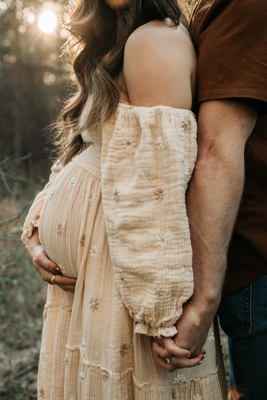 A pregnant woman and a man holding hands in a forest during sunset.