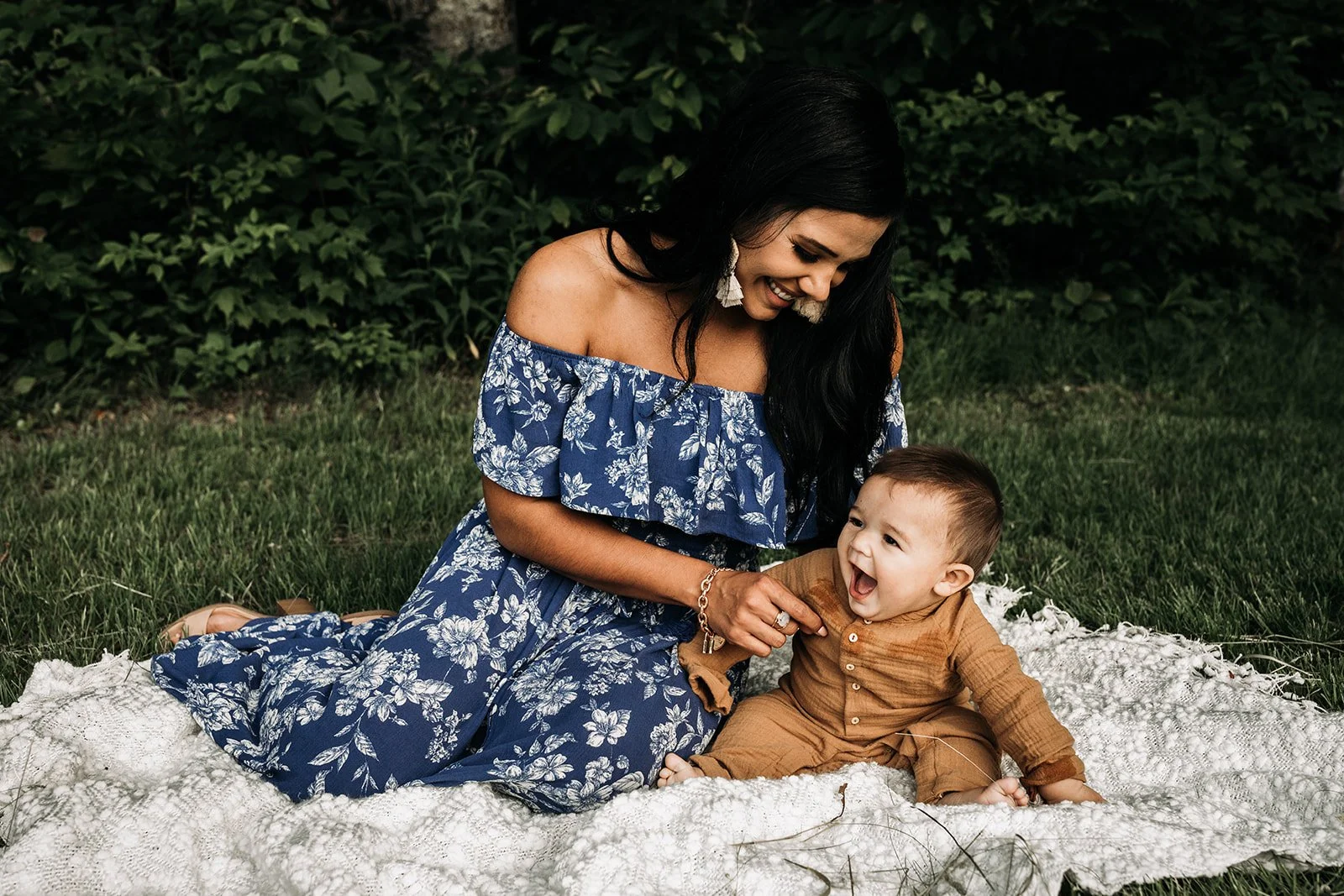A woman and a baby sitting on a white blanket outdoors, playing and smiling, with green bushes in the background.