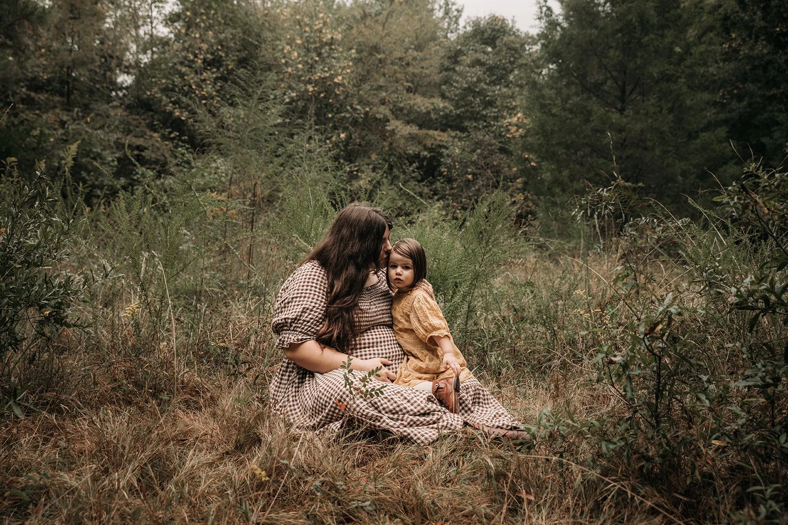 A woman and a young girl sitting on the grassy ground in a forested area, with the woman kissing the girl's forehead.
