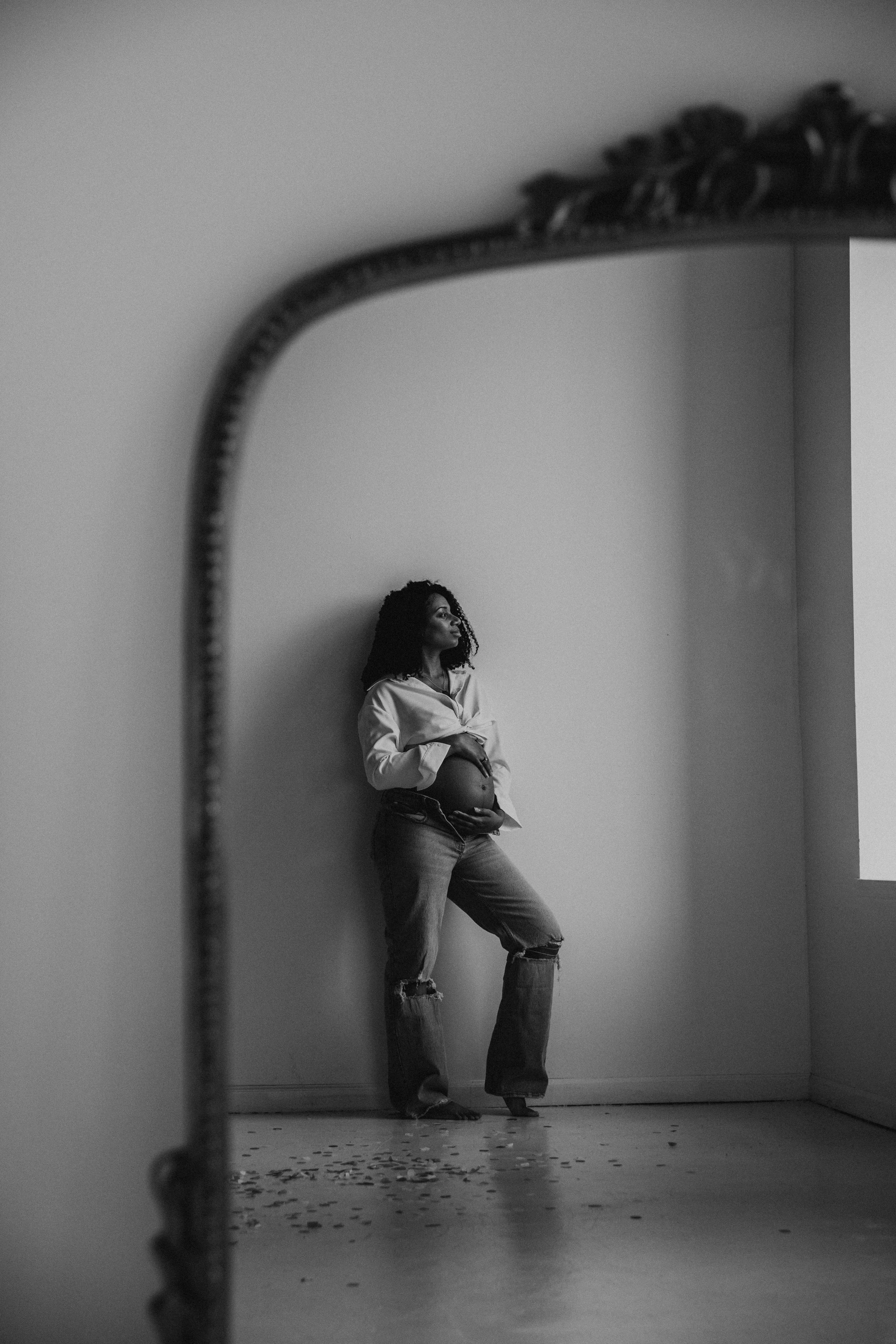 A pregnant woman stands by a window, looking away, reflected in a large mirror, in a minimalist room