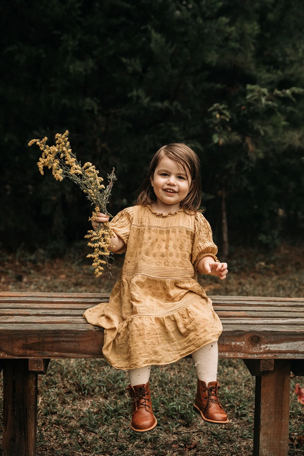 A young girl sitting on a wooden bench outdoors, holding a bouquet of yellow wildflowers, wearing a light brown dress and brown boots, smiling at the camera beside a wooded area.