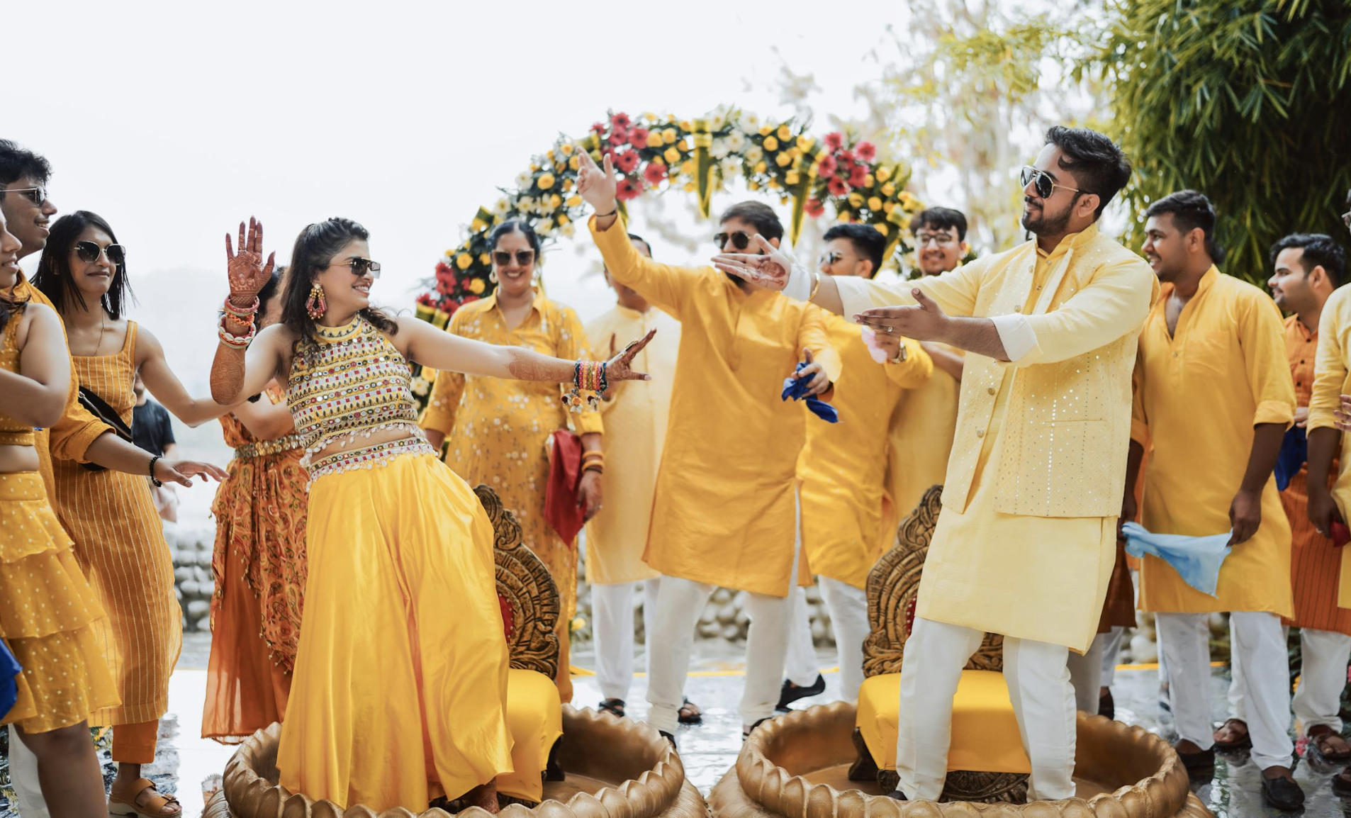 Group of people celebrating a traditional Indian wedding outdoors, wearing yellow and orange attire, dancing and smiling in front of a floral arch.