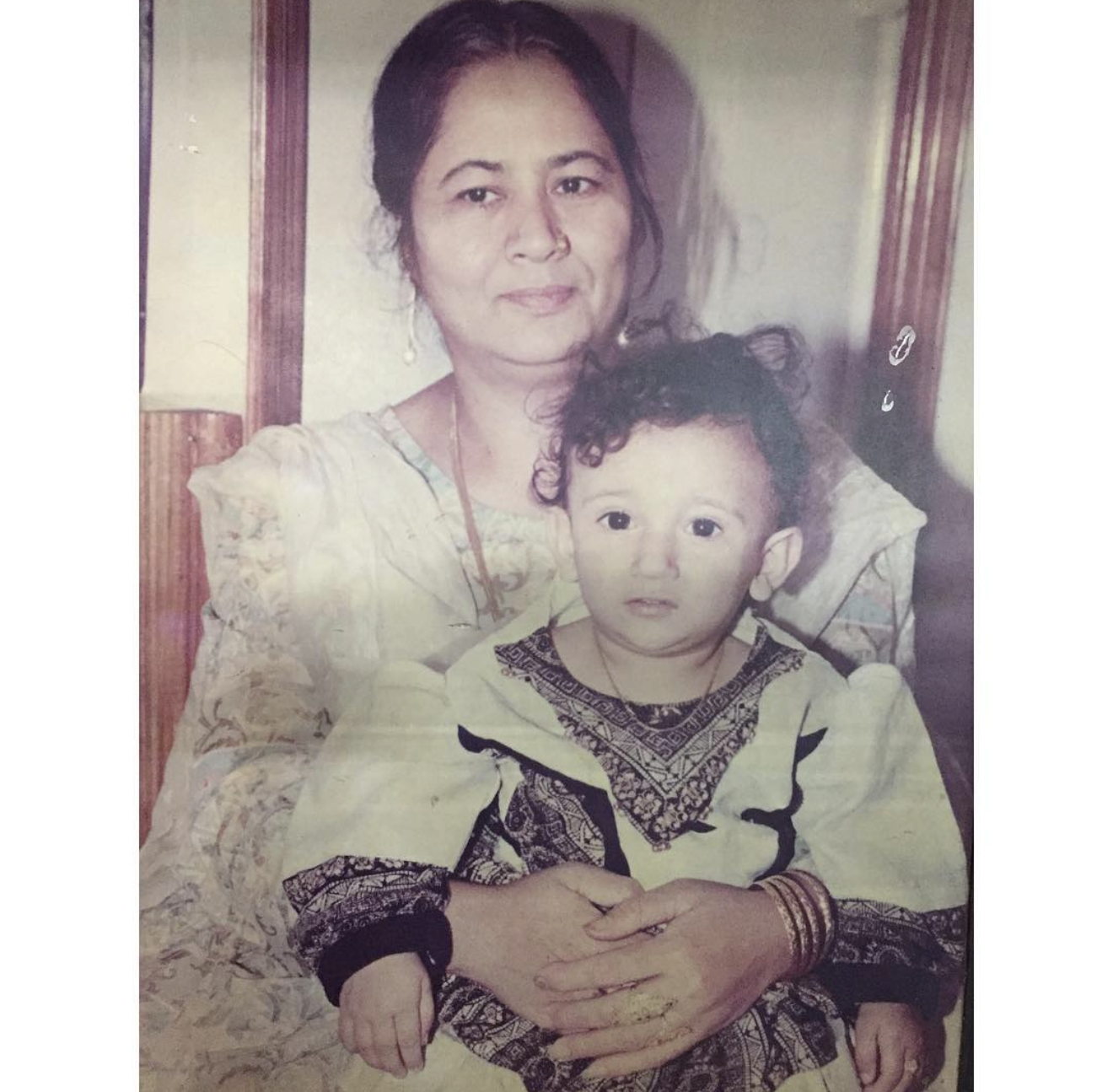 A woman holding a young child, both looking at the camera in a cozy indoor setting.
