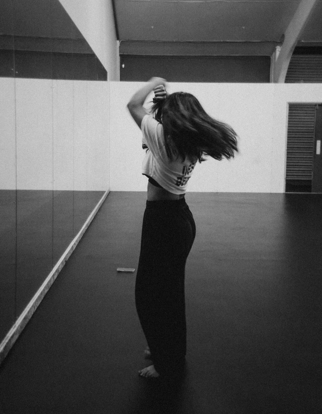 A woman with long hair standing on a dance studio floor, stretching her arms behind her head.