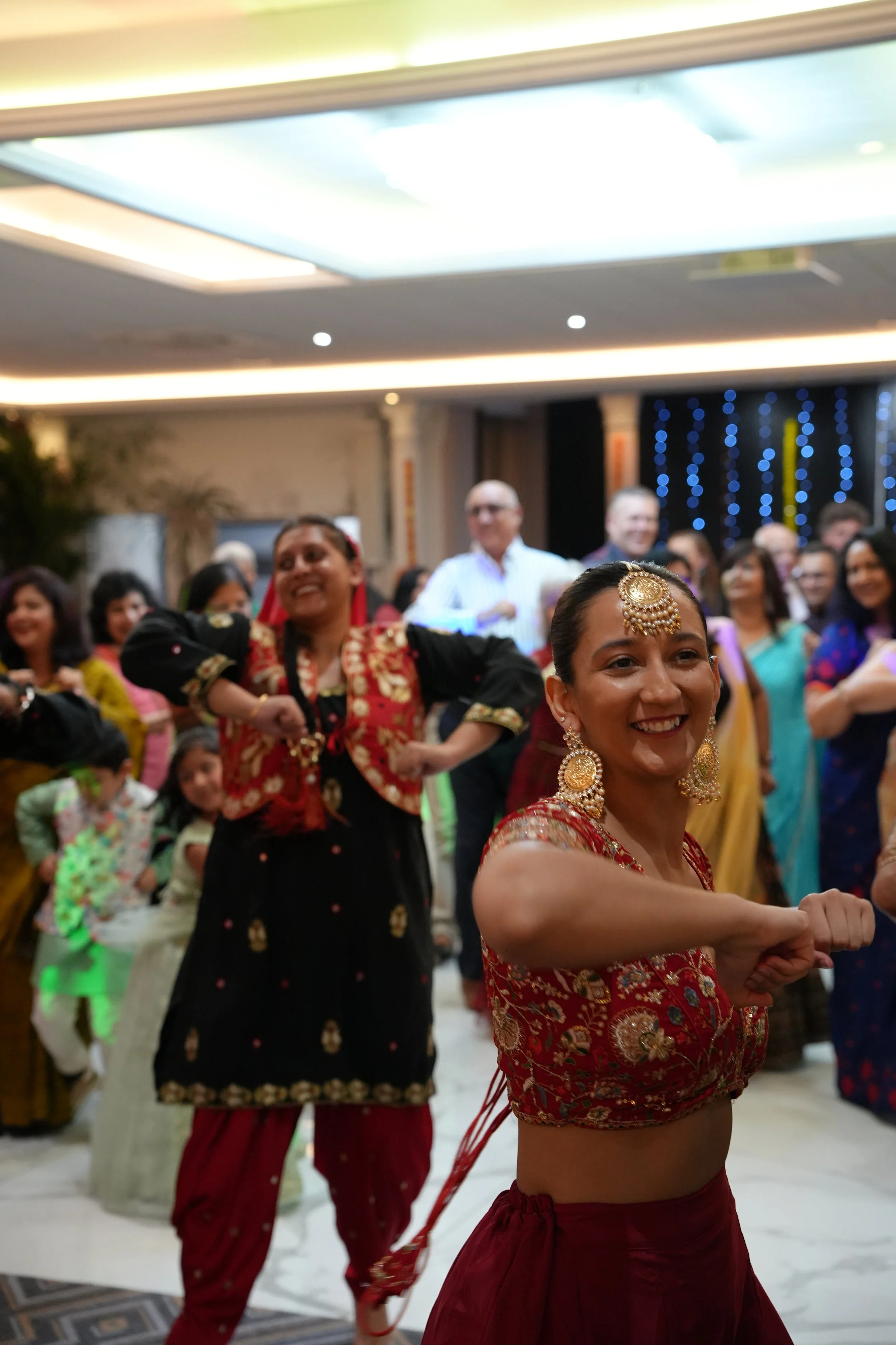 A group of women and children dancing at an indoor celebration, with one woman in the foreground wearing traditional Indian attire and jewelry.